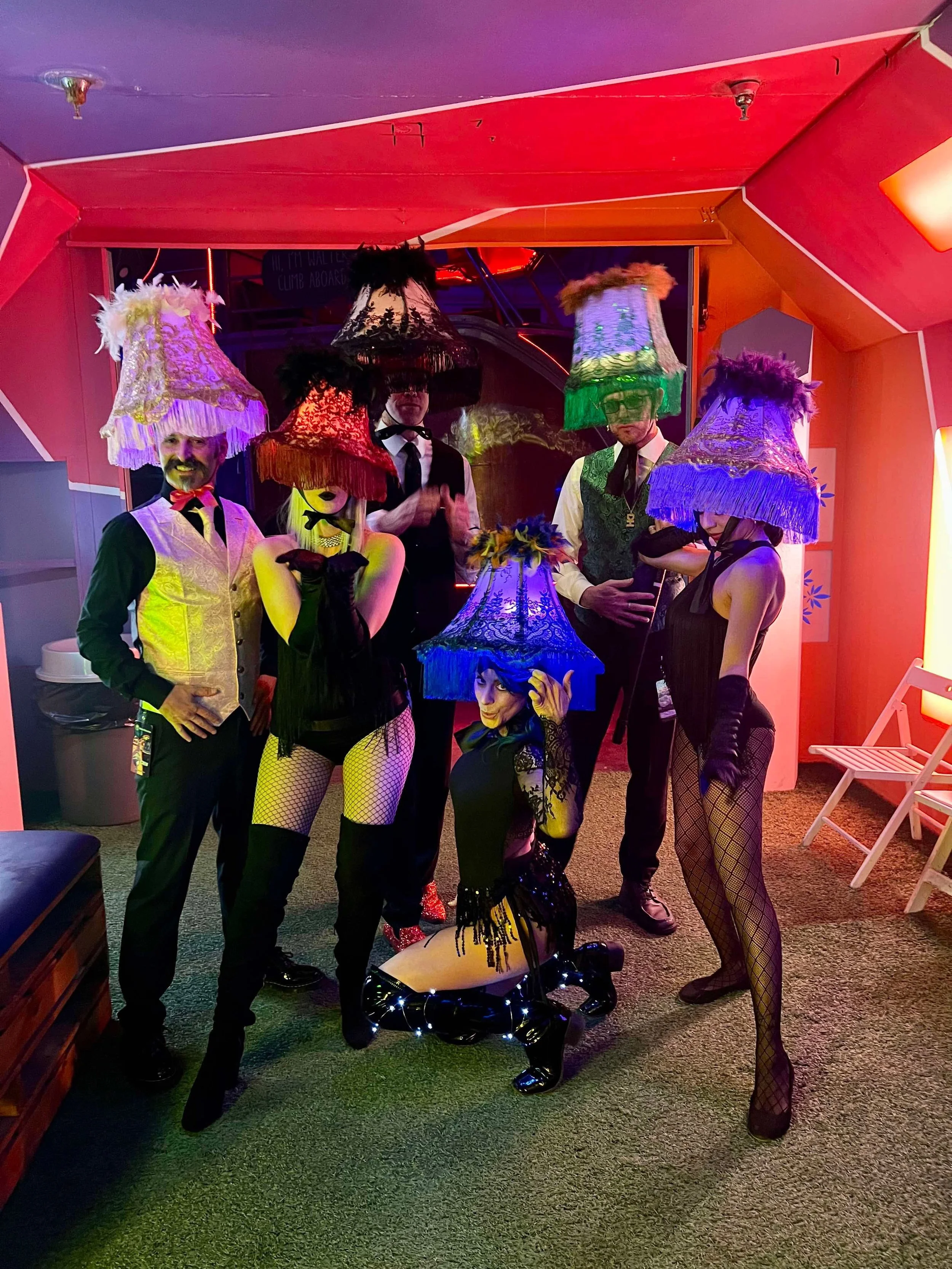 Group of people dressed in costumes with large, colorful lampshades on their heads, at a lively party or event in a room with red and purple lighting.