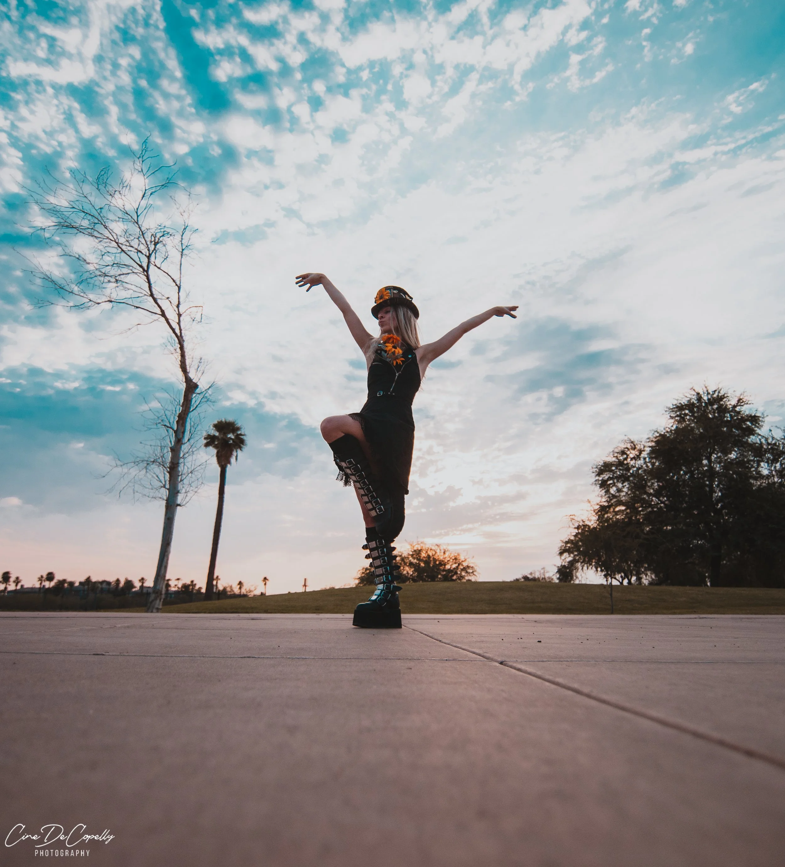 Person dancing outdoors at sunset, wearing platform boots, a black outfit, and a headband, with trees and a partly cloudy sky in the background.