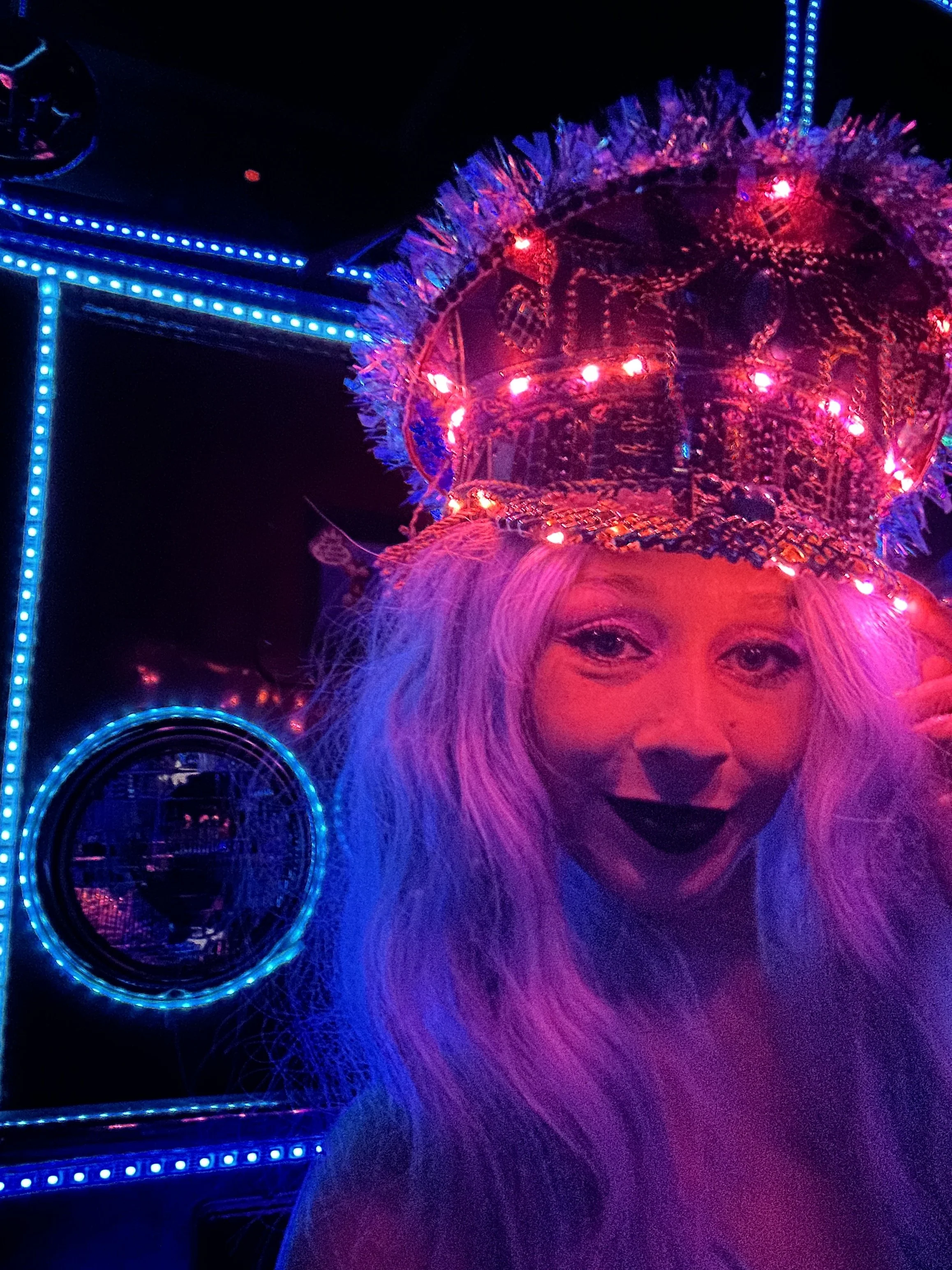 A blonde woman wearing a detailed, brightly lit, crown with pink lights. black lipstick.The background is decorated with colorful LED lights and reflective surfaces. captain hat