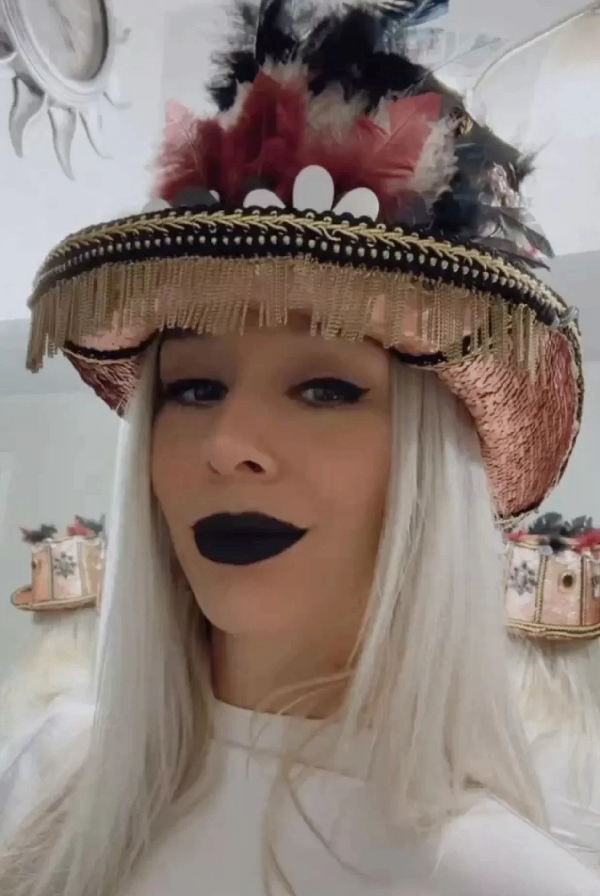 A woman with long platinum blonde hair wearing an ornate, vintage-style top hat decorated with feathers, flowers, and fabric details, and black lipstick.
