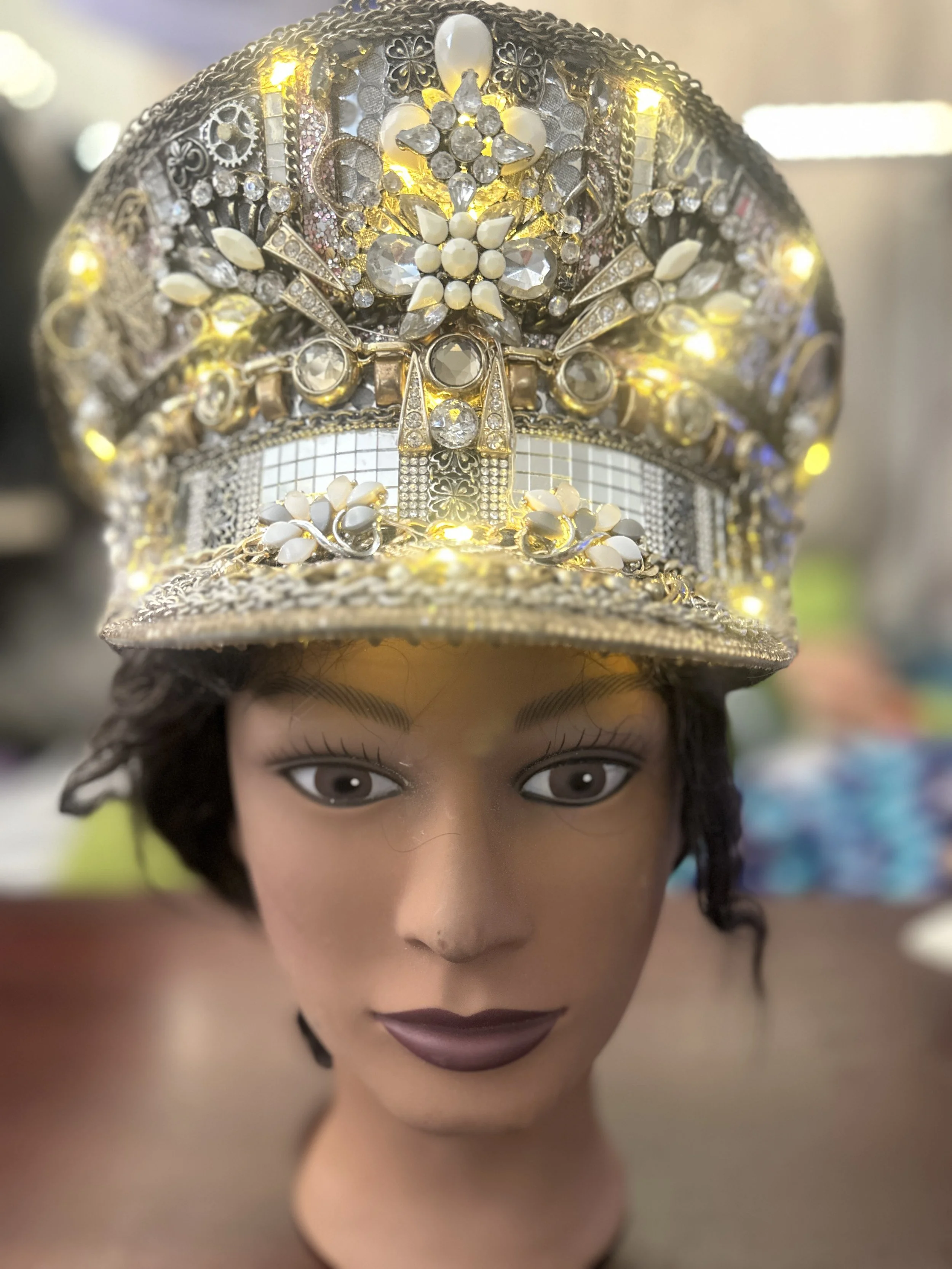 A mannequin head wearing a silver, jeweled, illuminated hat decorated with rhinestones, flowers, and intricate metalwork.