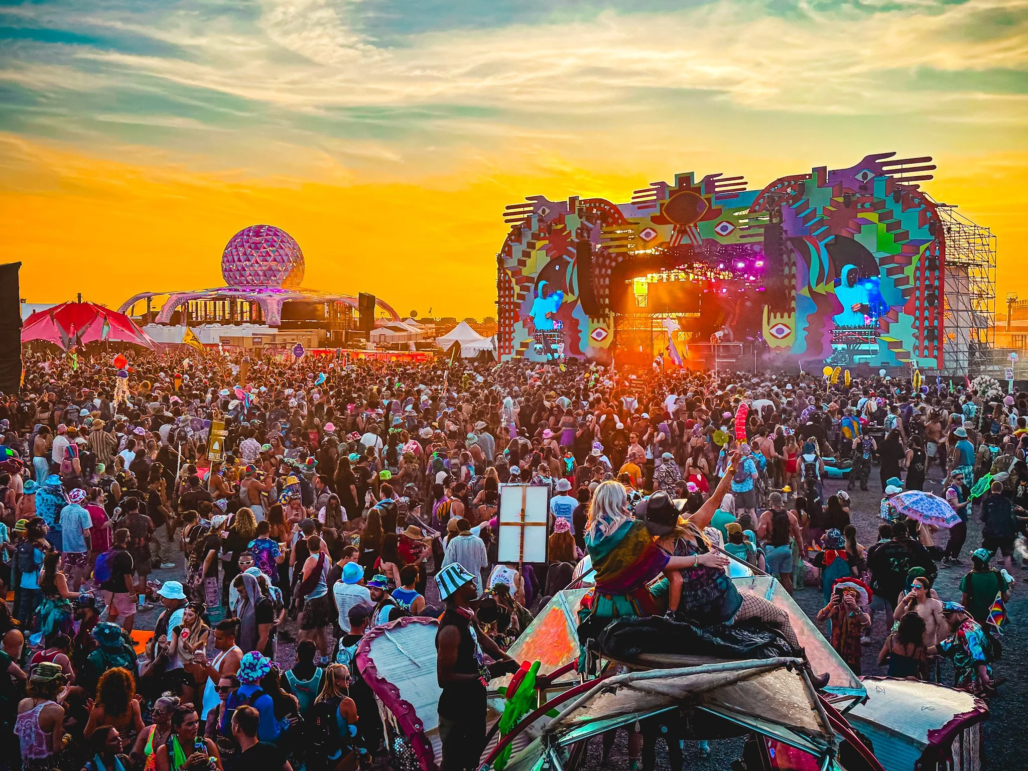 Crowd of people at a music festival during sunset with a colorful stage and a large spherical structure in the background.