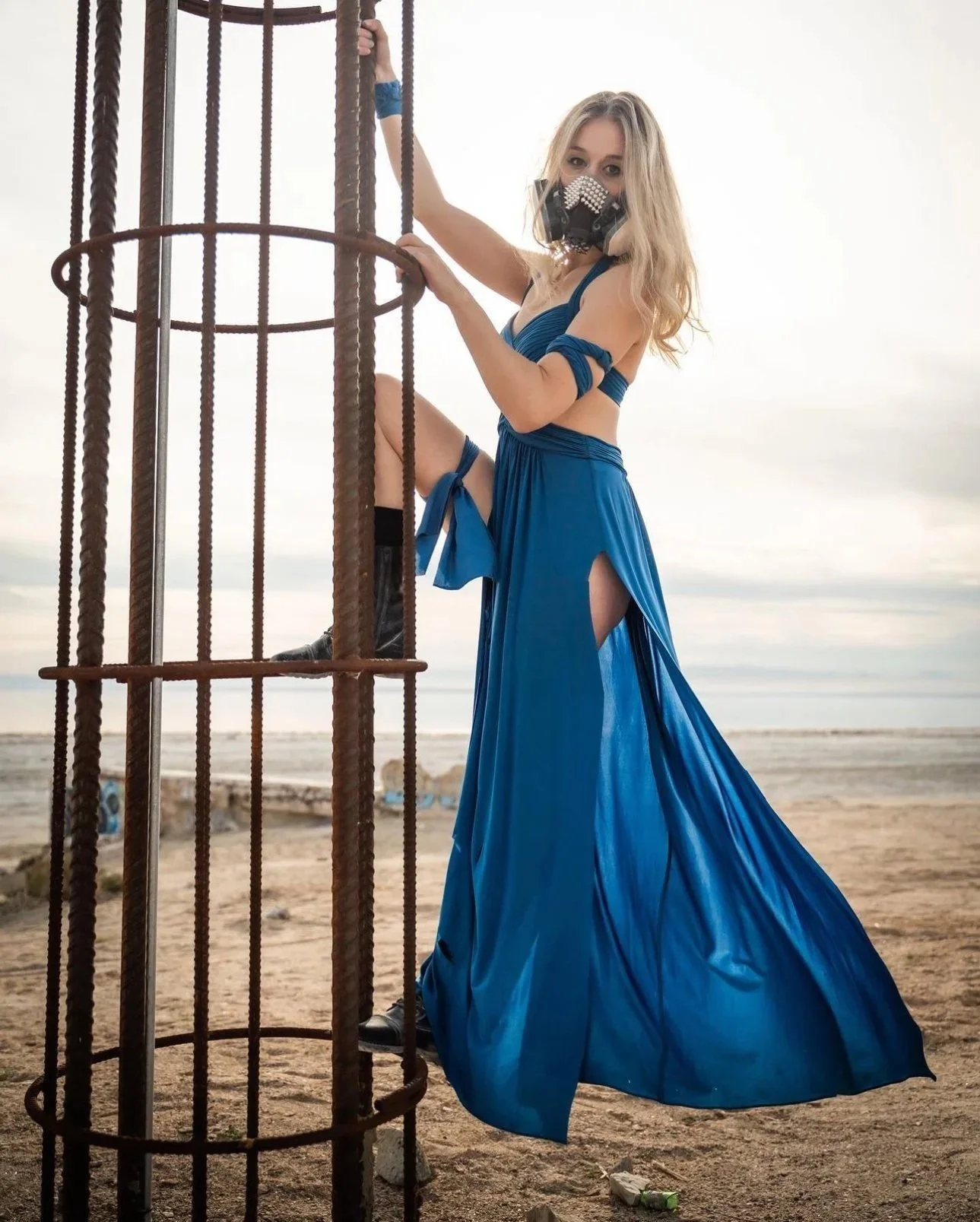 Blonde Woman in a blue dress and a gas mask climbing a metal structure on a beach.