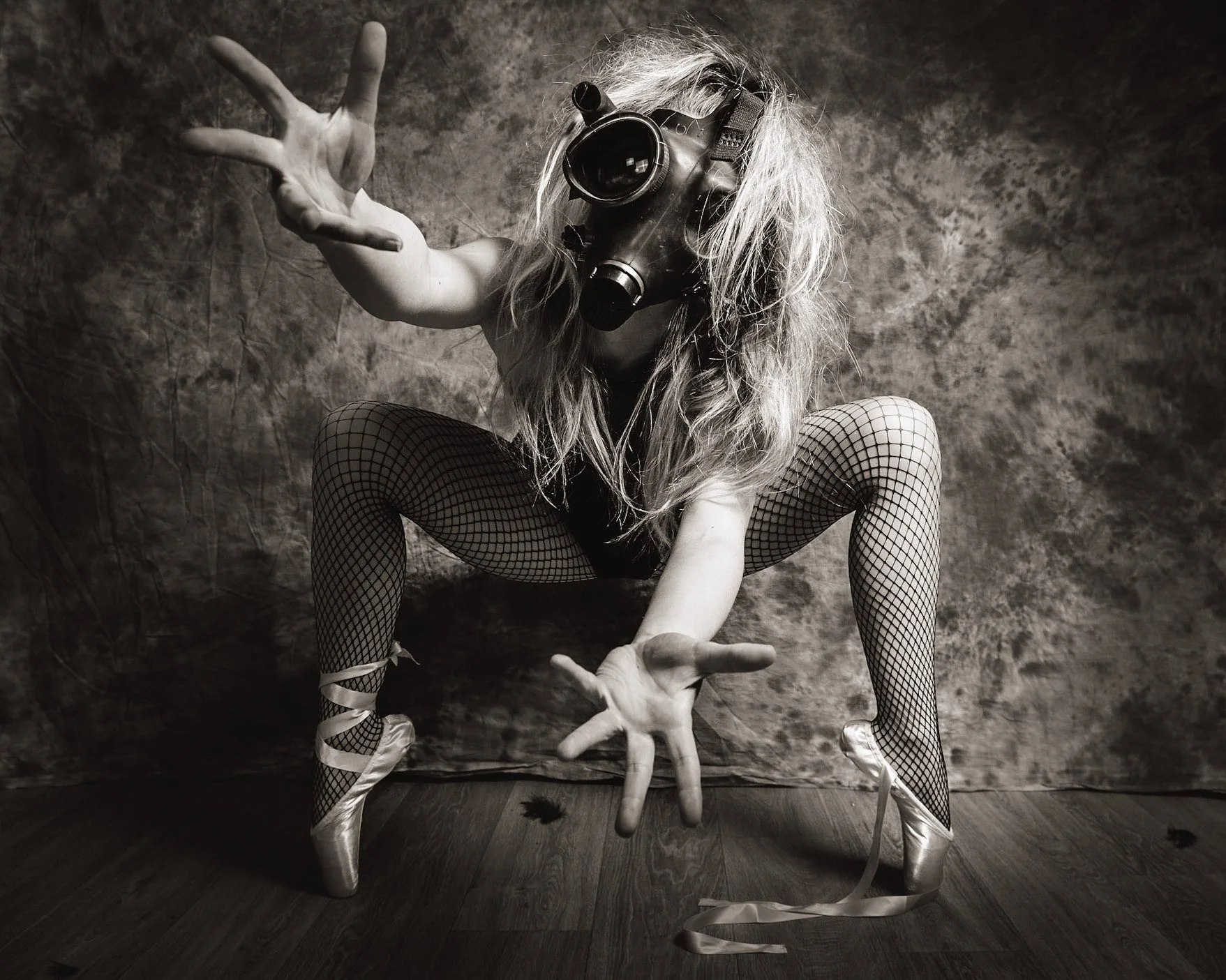 A person wearing a gas mask, fishnet stockings, ballet pointe shoes, and a wig, posed in a squatting position with arms extended forward and fingers spread, against a textured backdrop in a black-and-white photograph.