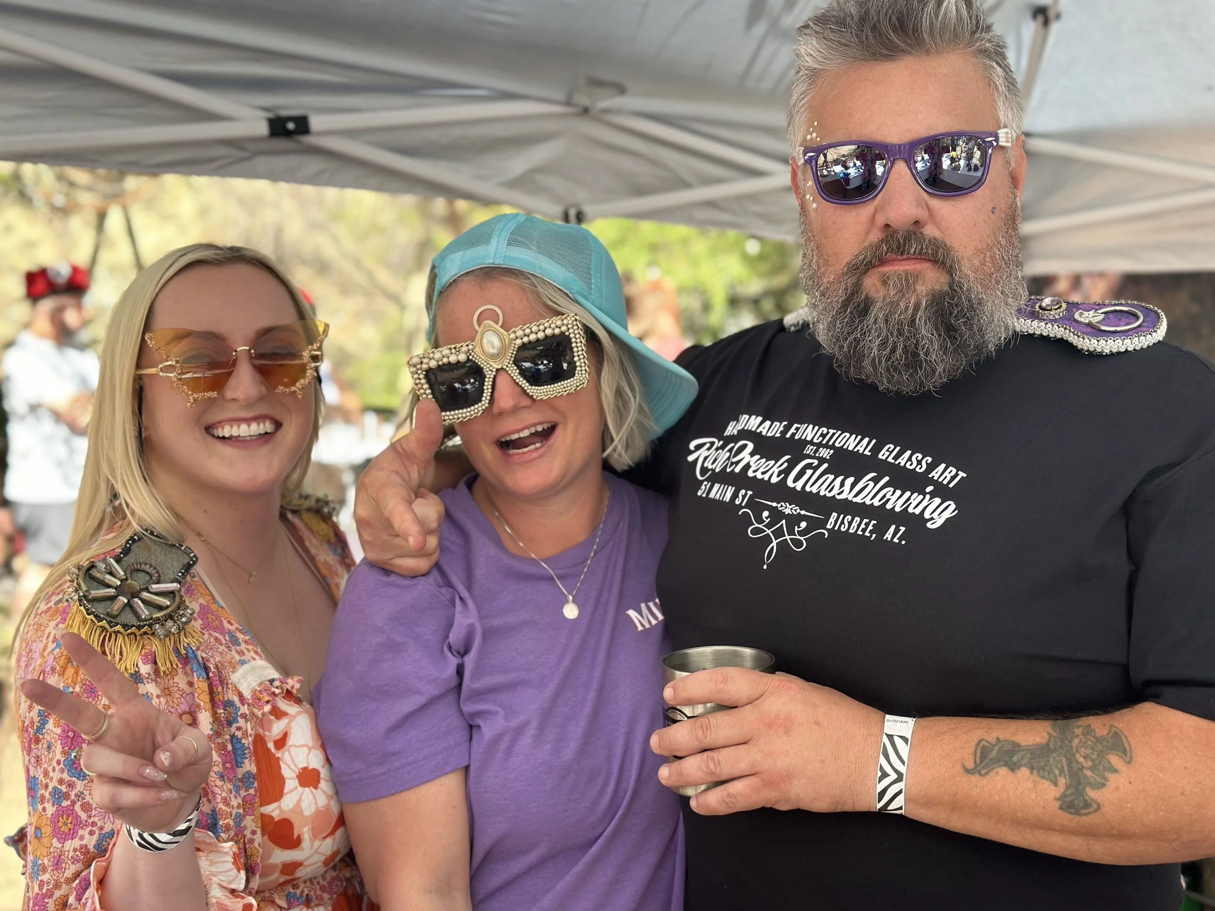 Three people smiling and posing at an outdoor event, wearing sunglasses and colorful clothing, under a tent with trees in the background.