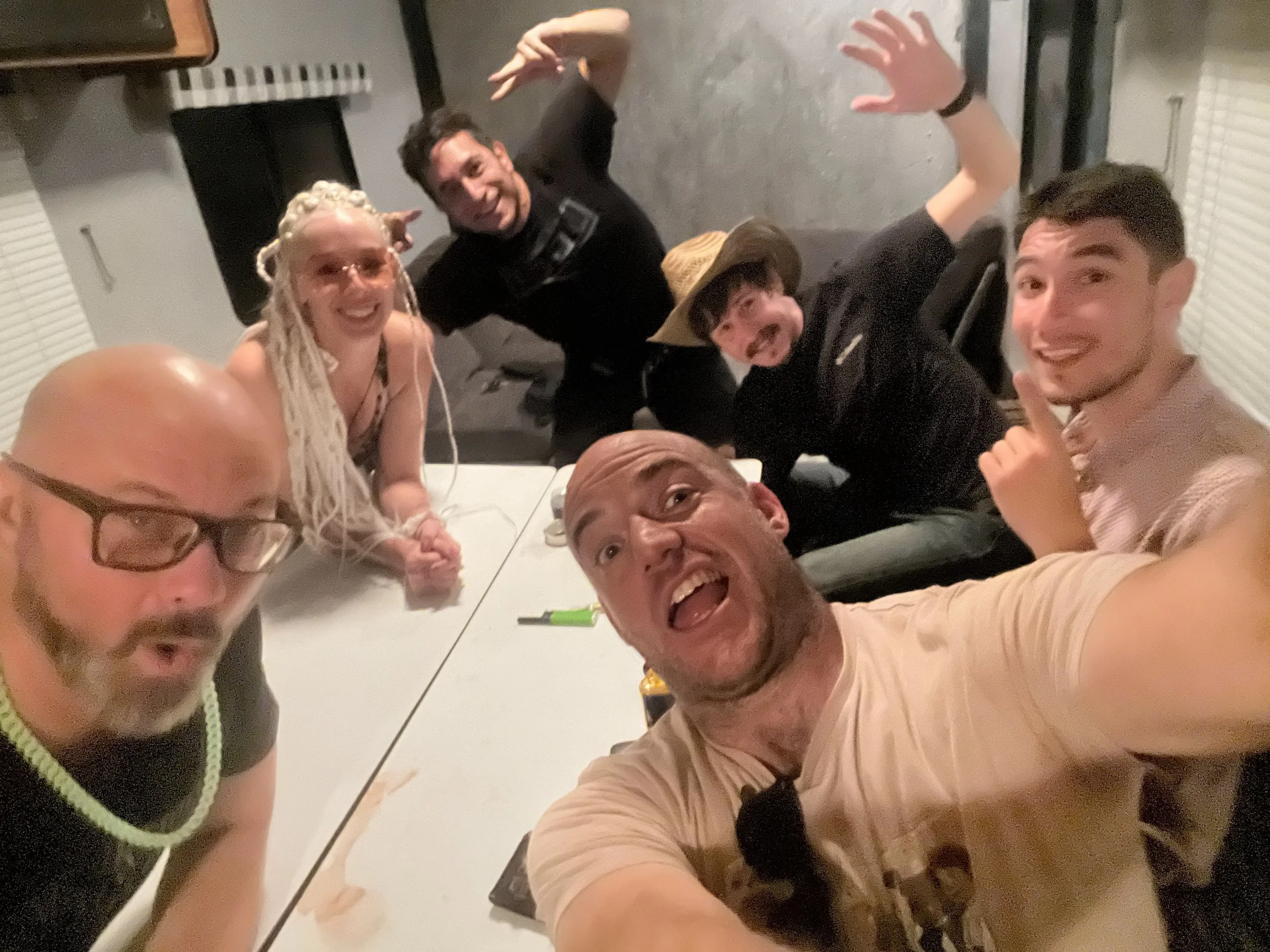 Group of seven people enjoying a party, taking a selfie together at a table in a room with blinds.