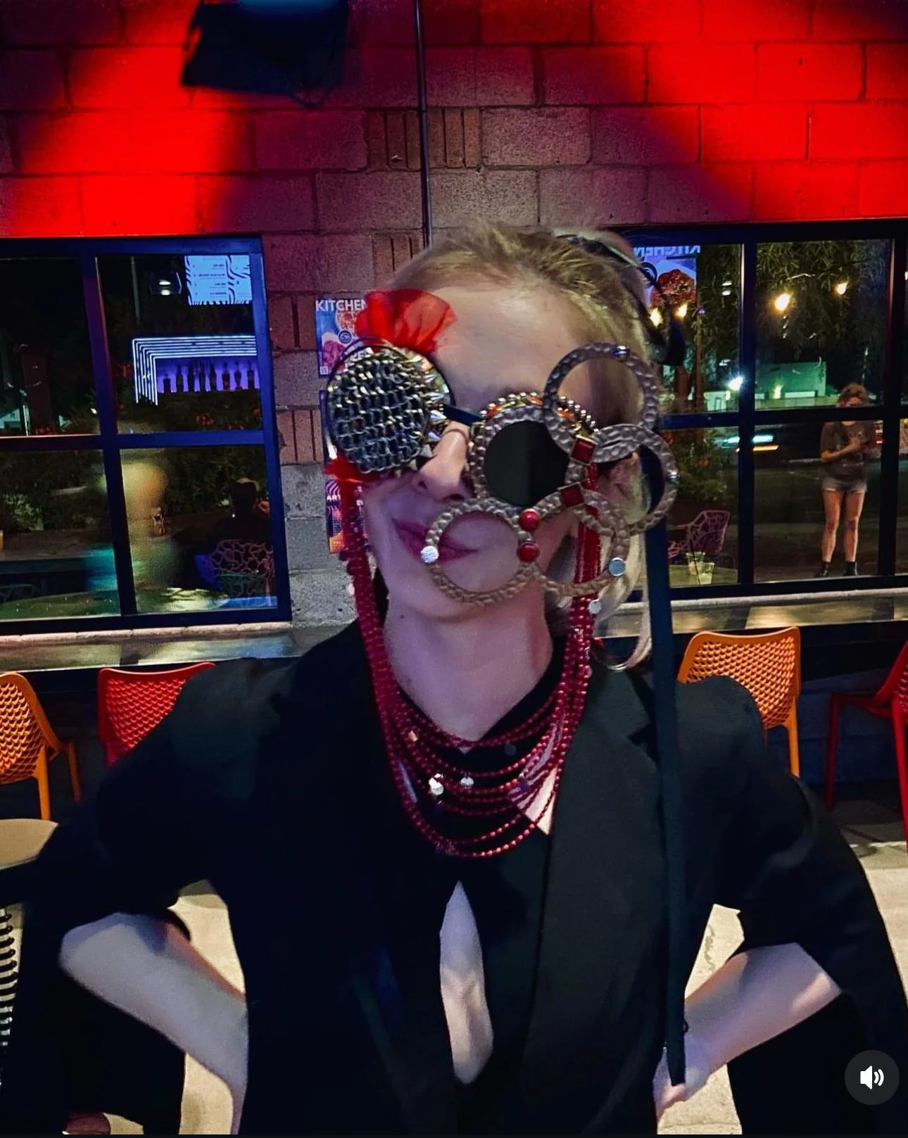 A woman in a black top with red jewelry and wearing multiple large, decorative earrings and sunglasses, posing with her hands on her hips in a colorful indoor setting with large windows, red lighting, and a view of the outdoors.