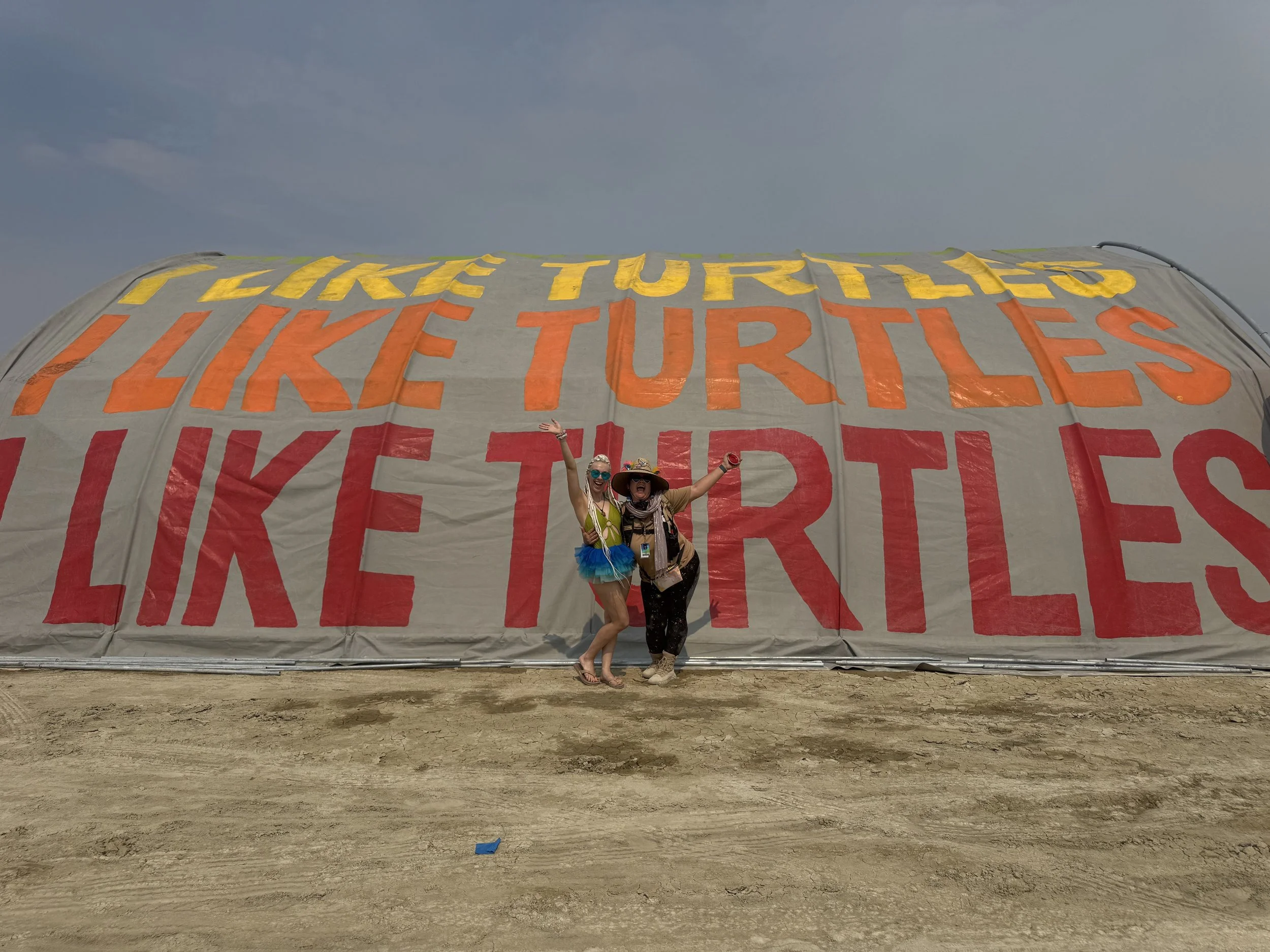 Two women standing in front of a large tent with colorful text reading "Like Turtles". They are celebrating, smiling, with arms raised, in a desert setting under a cloudy sky.