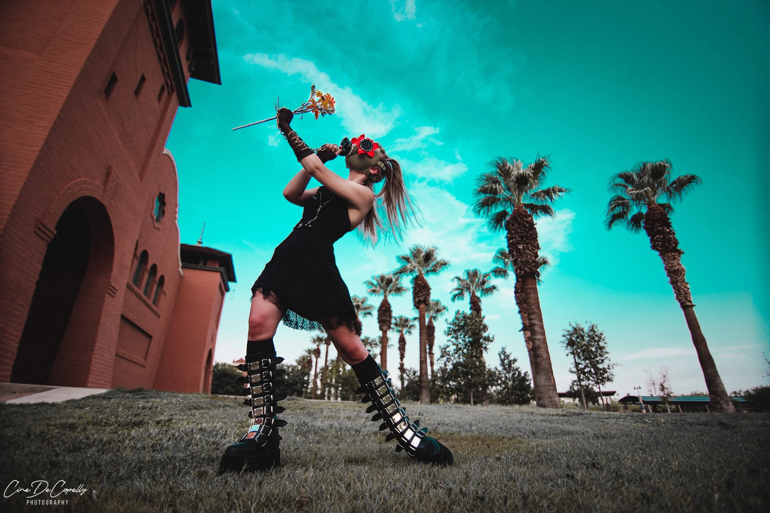 A woman with long blonde hair and a colorful headband, wearing a black dress with mesh accents, stands on grass holding a flower to her lips. She is wearing tall, black, multi-buckle platform boots, and has black gloves on her arms. The background features a red brick building, tall palm trees, and a bright blue sky with some clouds.