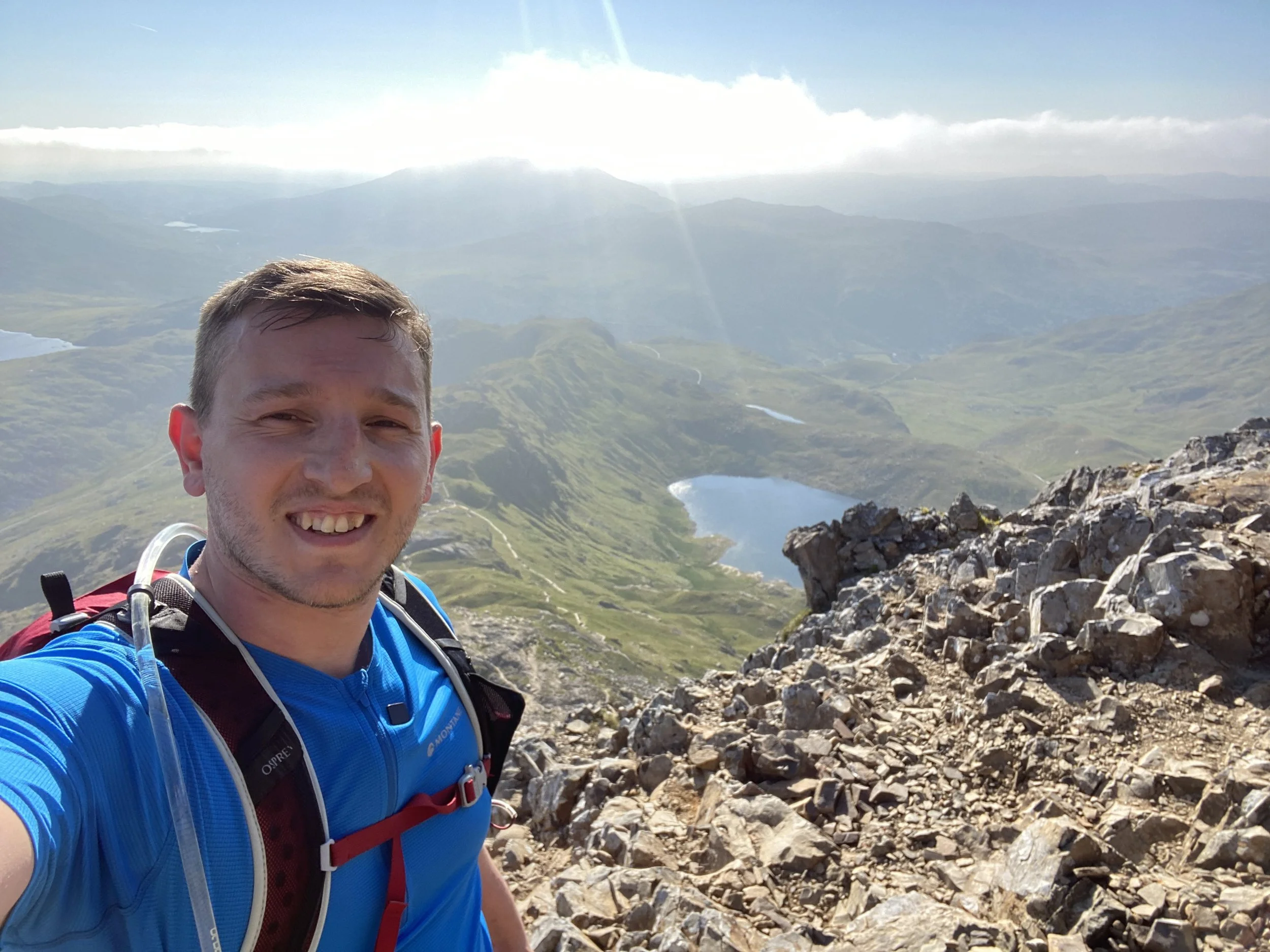 A man with short hair and a beard taking a selfie on a rocky mountain trail, with a scenic view of lakes and green hills and mountains in the background.