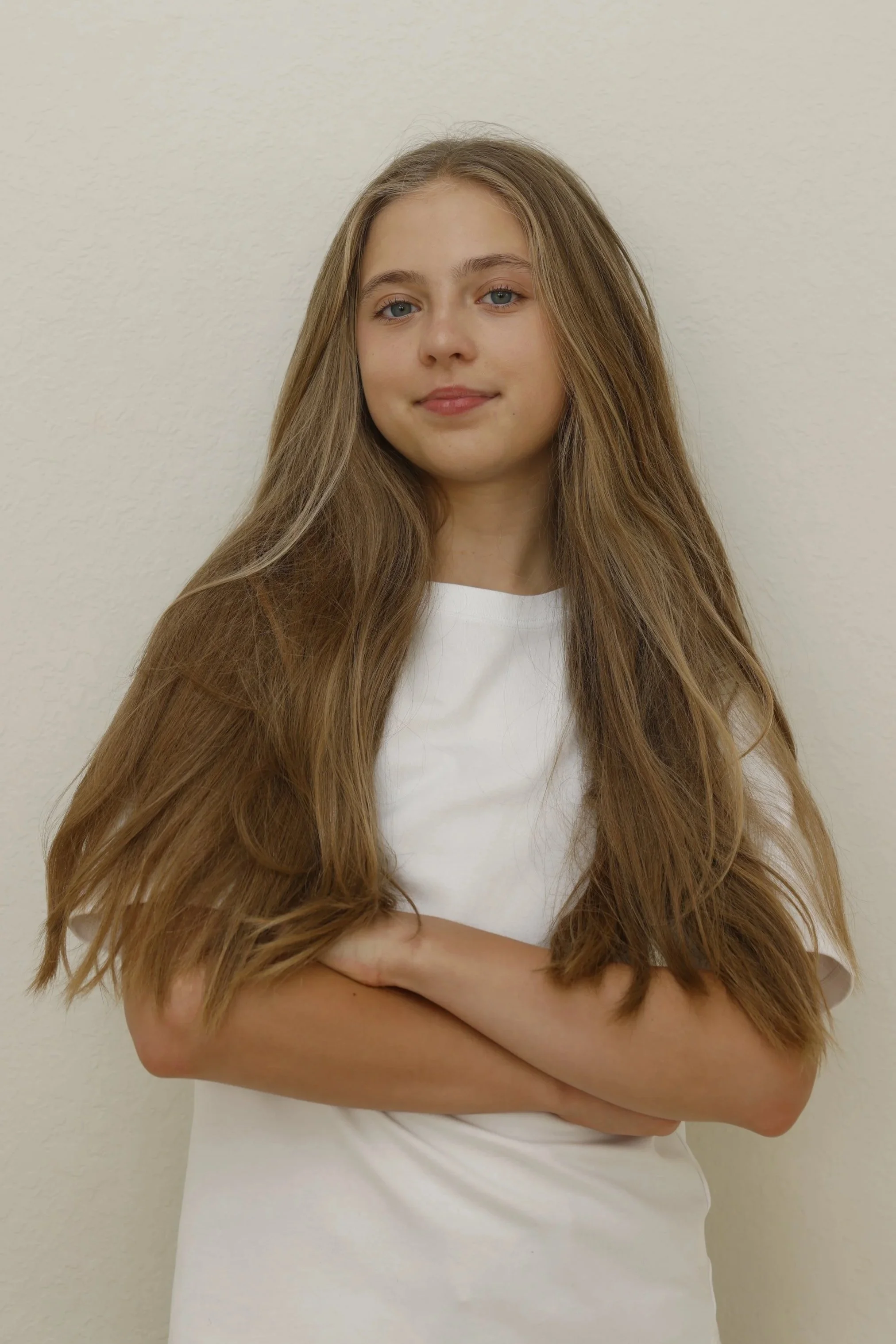 A young woman with long, wavy light brown hair and blue eyes, standing against a plain beige wall, wearing a white T-shirt and crossing her arms.