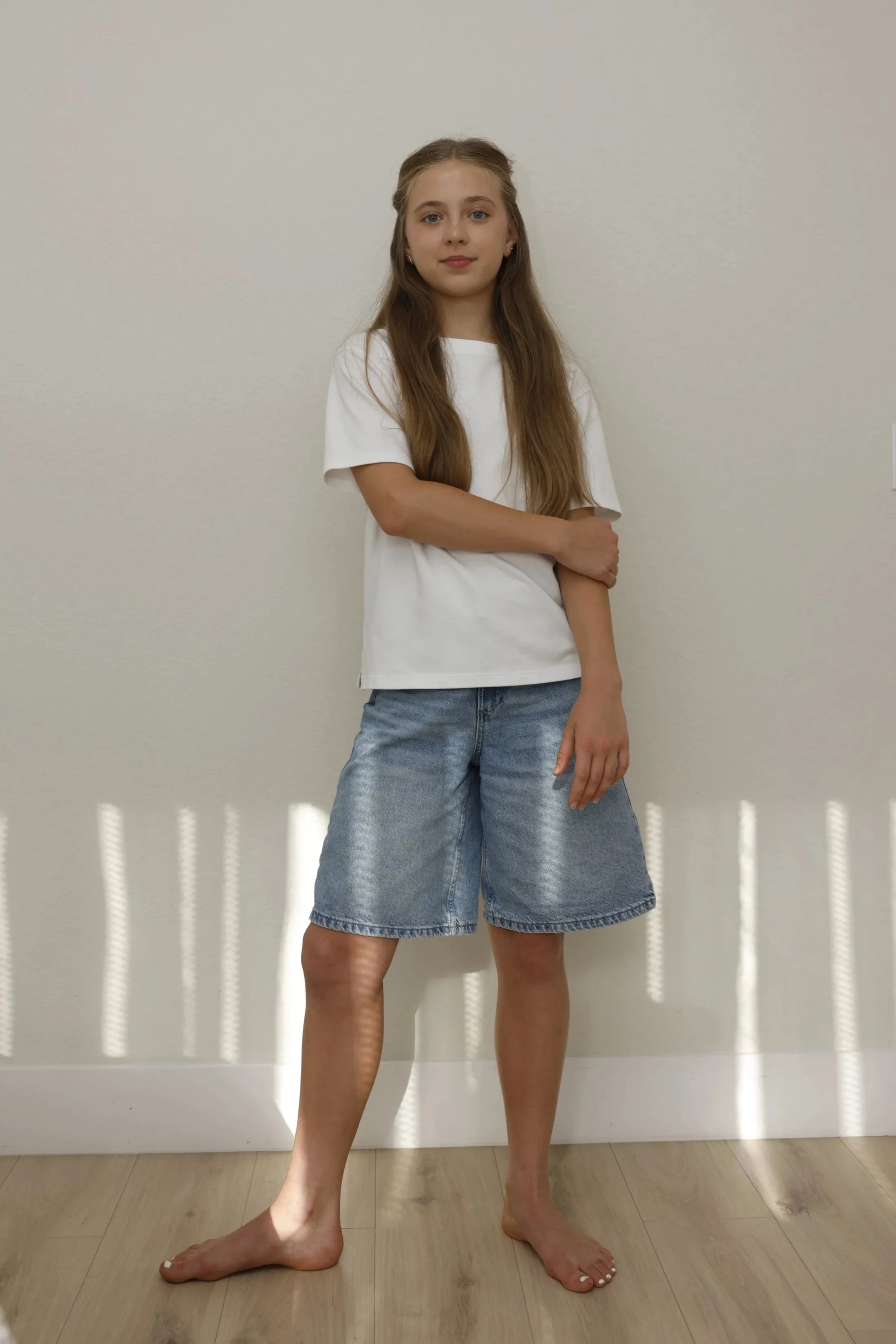 A young girl with long brown hair and blue eyes standing barefoot against a plain white wall, wearing a white t-shirt and denim shorts, with her arms crossed in front of her.