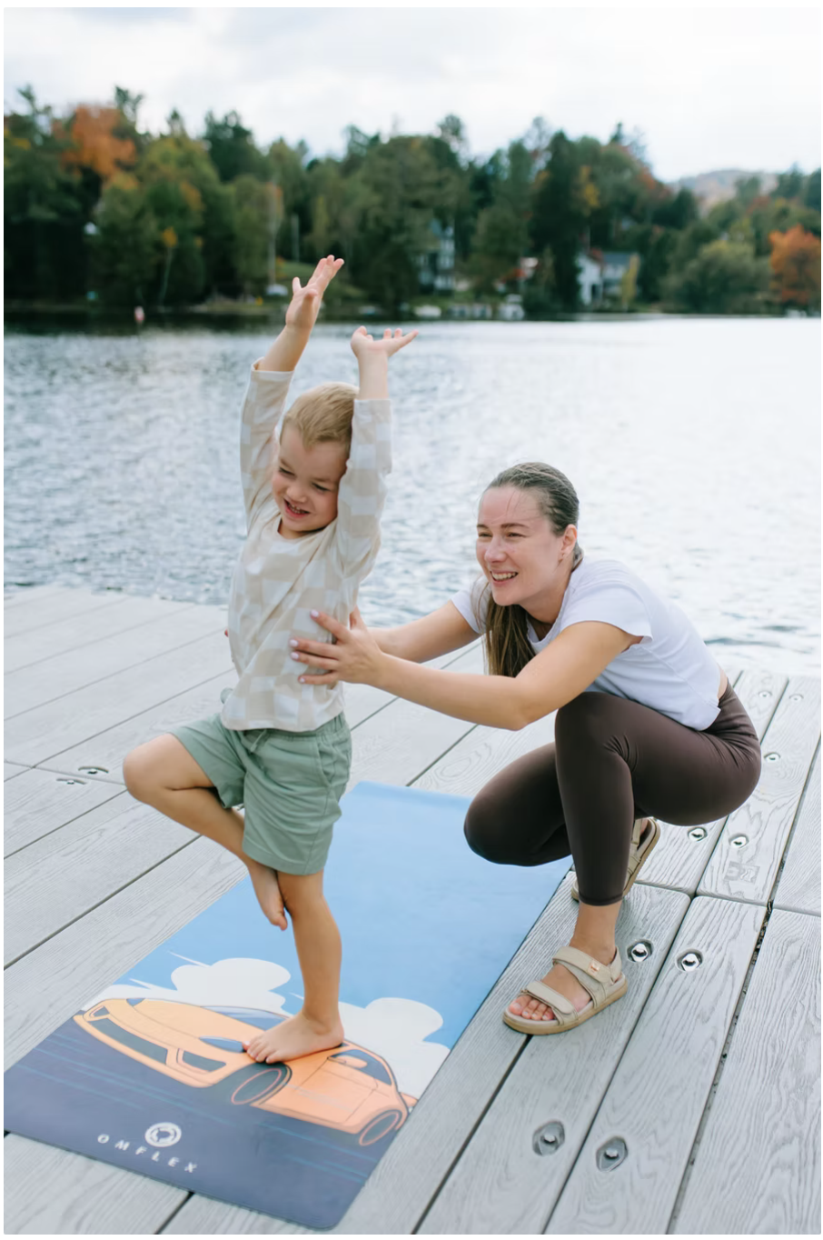 A woman helping a young boy do yoga on a dock by a lake during autumn, with trees and houses in the background.