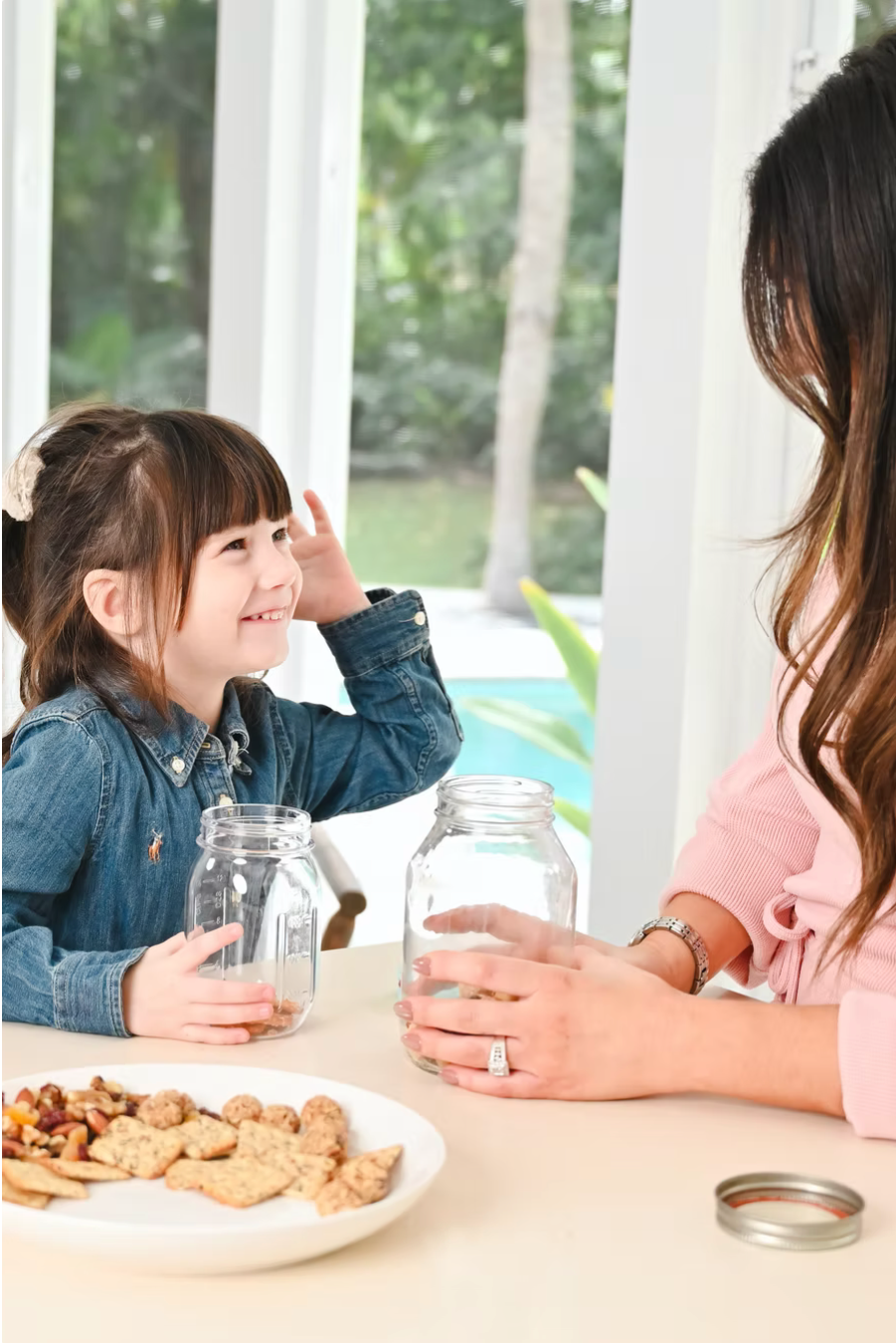 A young girl and woman sitting at a table with jars, a plate of cookies and snacks, by large windows showing trees outside. The girl is smiling and listening to the woman.