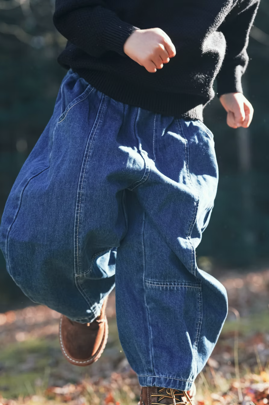 Close-up of a child running outdoors, wearing baggy blue jeans, a black sweater, and brown boots, with a background of blurred nature.