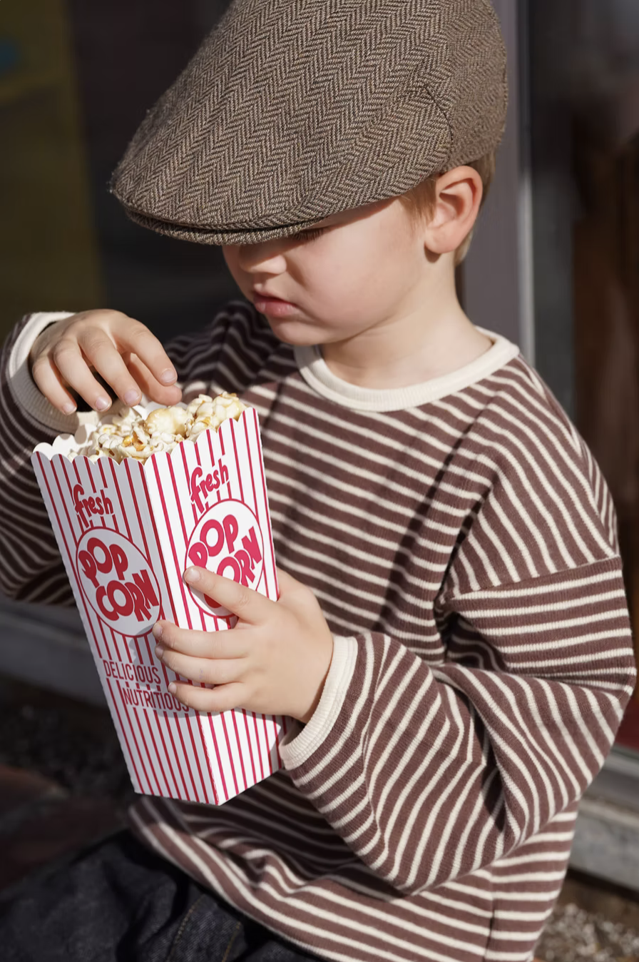 A young boy wearing a brown striped shirt and a brown hat holding a large container of popcorn.