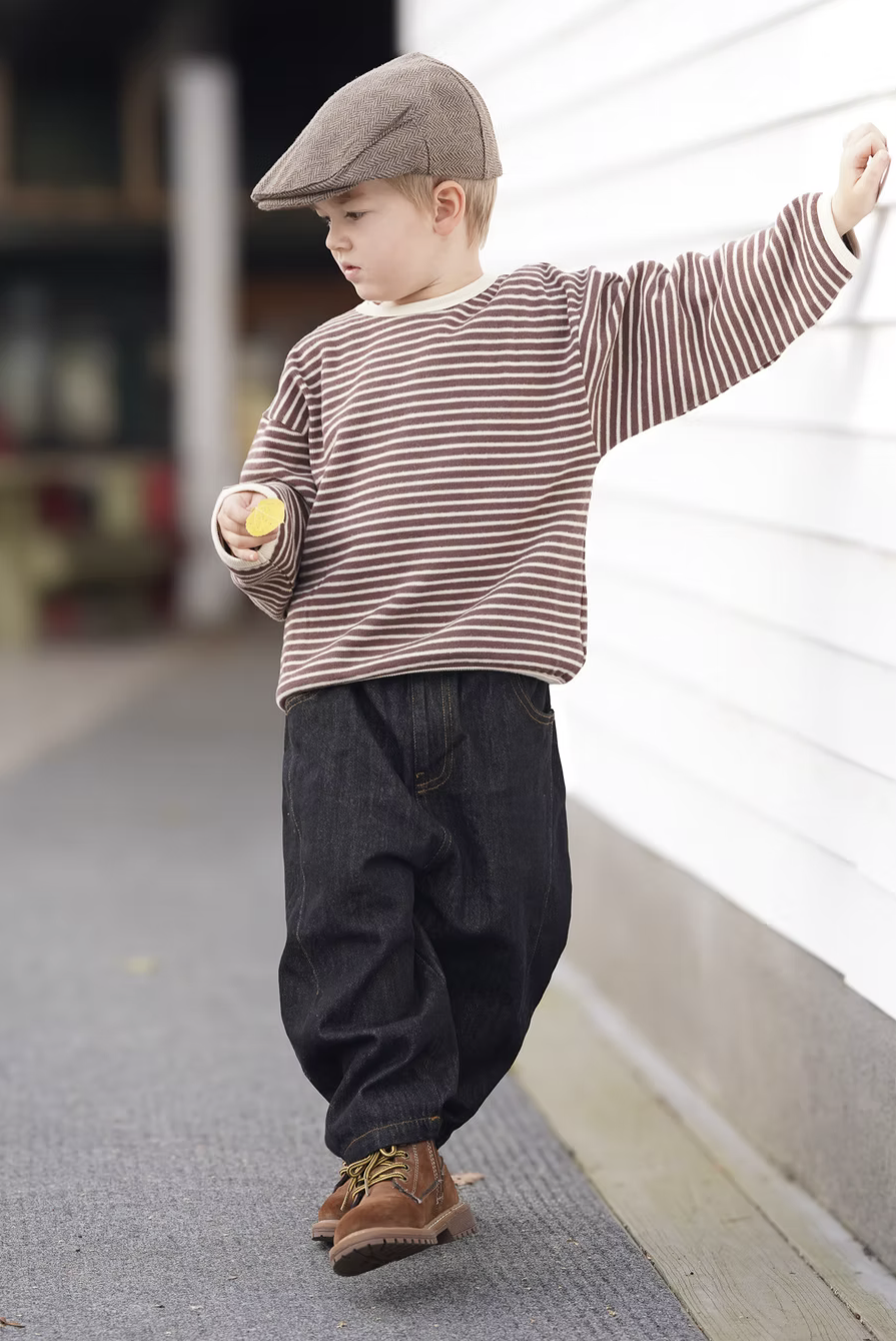 A young boy dressed in a striped long sleeve shirt, dark jeans, brown boots, and a flat cap, walking along a sidewalk while holding a yellow object.