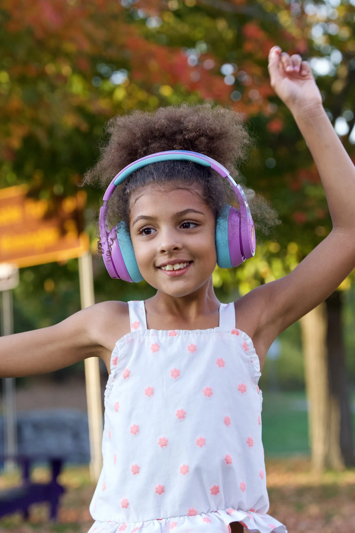 A young girl wearing purple and teal headphones, smiling, with her arm raised, outdoors in a park during fall.