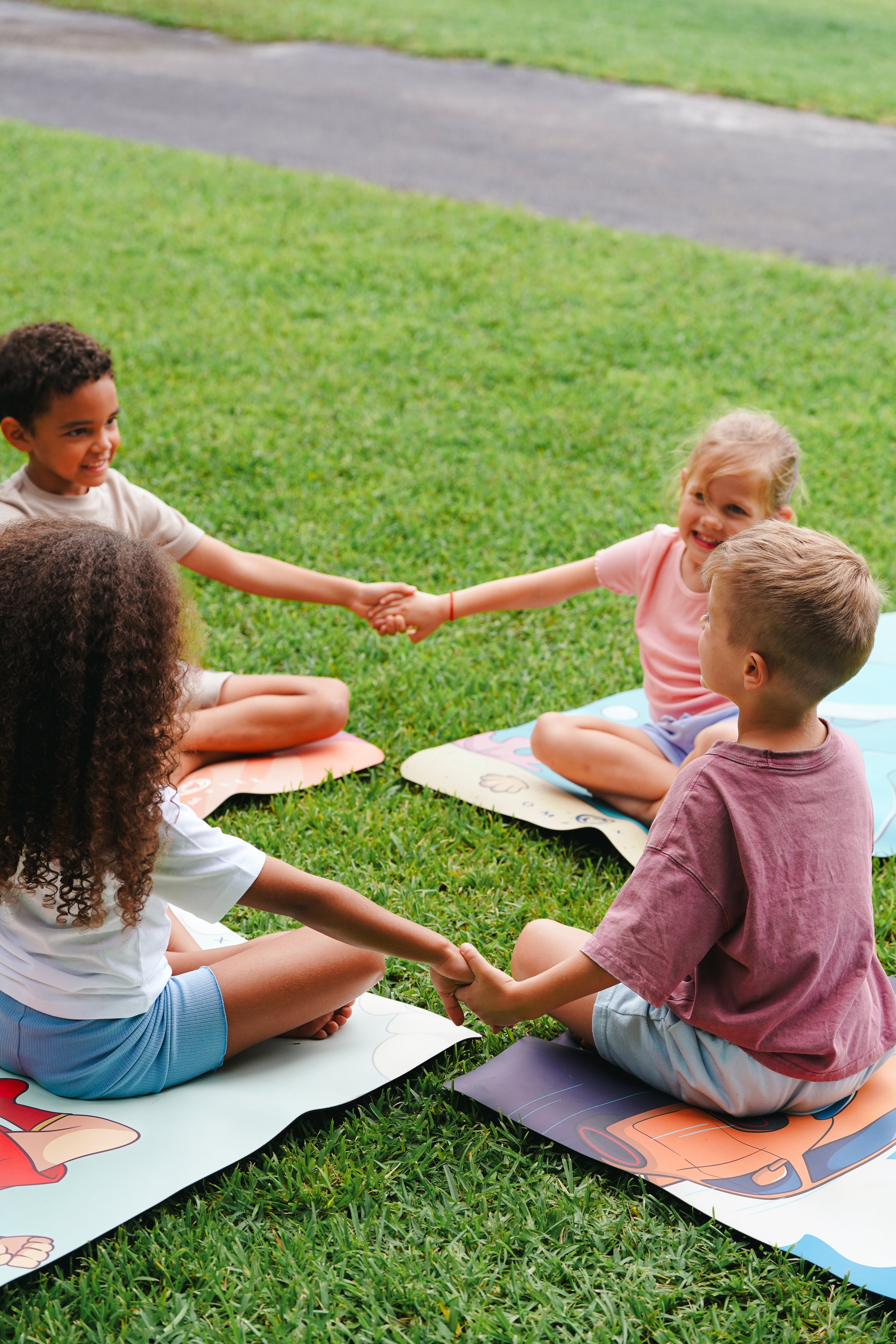 Children sitting on mats in a circle, holding hands, outside on a grassy area.