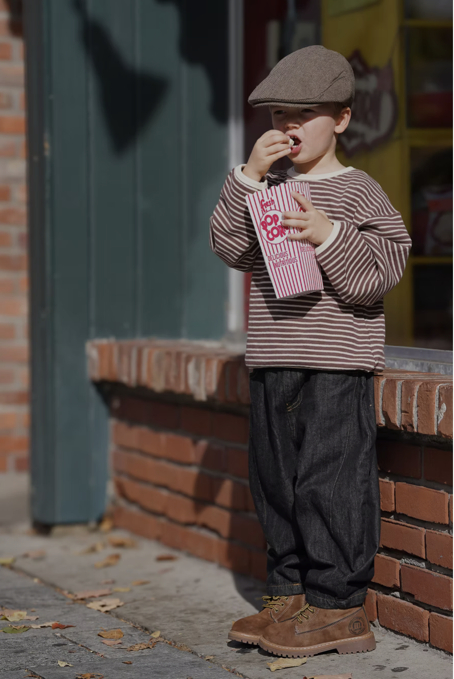 A young boy wearing a flat cap, striped long-sleeve shirt, dark jeans, and tan boots, eating popcorn outside a building on a sidewalk.