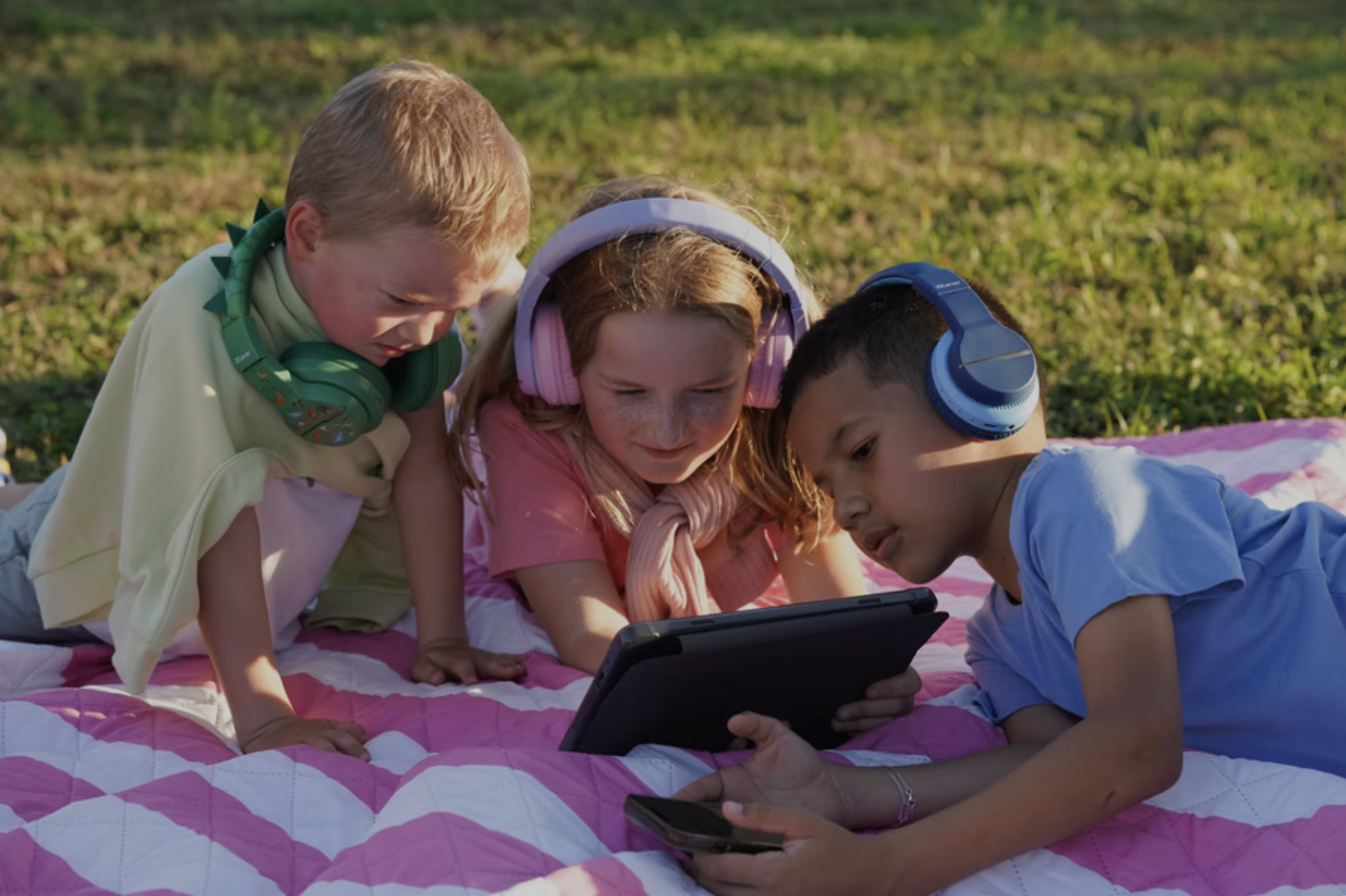 Three children lying on a pink and white blanket outdoors, wearing headphones, gathered around a handheld gaming device and a smartphone, looking at the device.