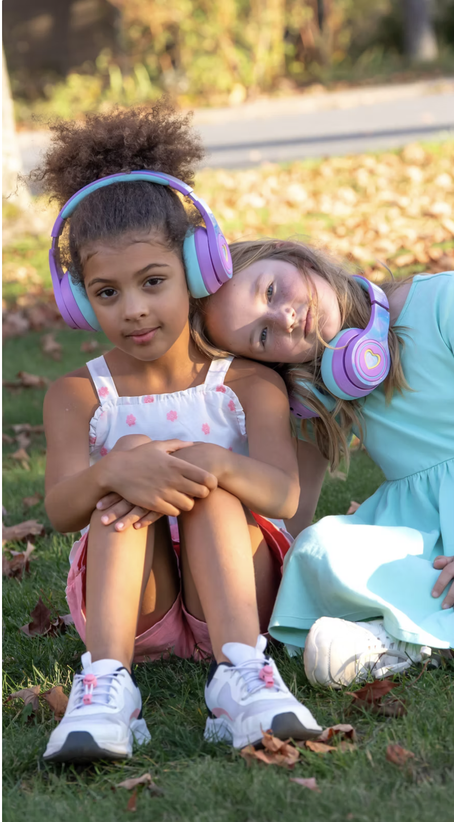 Two young girls sitting on grass outdoors, wearing large purple and turquoise headphones. One girl with curly hair in a white top with pink flowers, pink shorts, and white sneakers sitting with her knees up, and the other girl with straight hair in a blue dress, leaning her head on the first girl's shoulder. Autumn leaves are scattered on the ground.