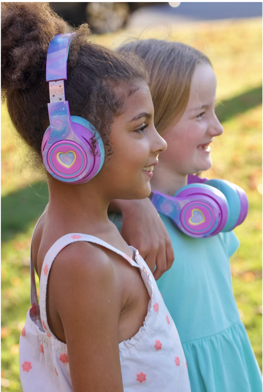 Two young girls smiling outdoors, wearing colorful headphones, one with curly hair and one with straight hair.