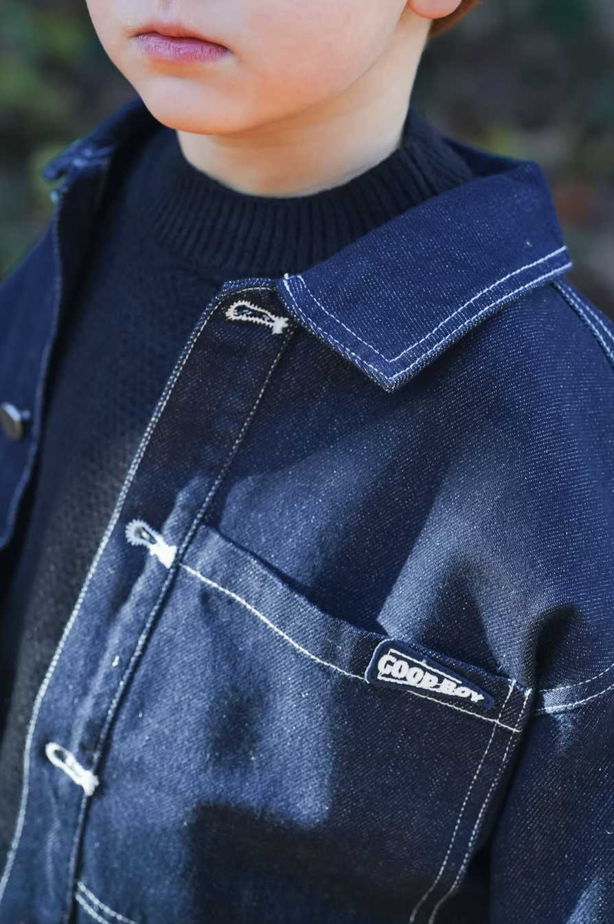 Close-up of a young boy wearing a dark blue denim jacket with white stitching and a small 'Good Boy' label on the chest pocket, over a black shirt, with part of his face and lips visible.