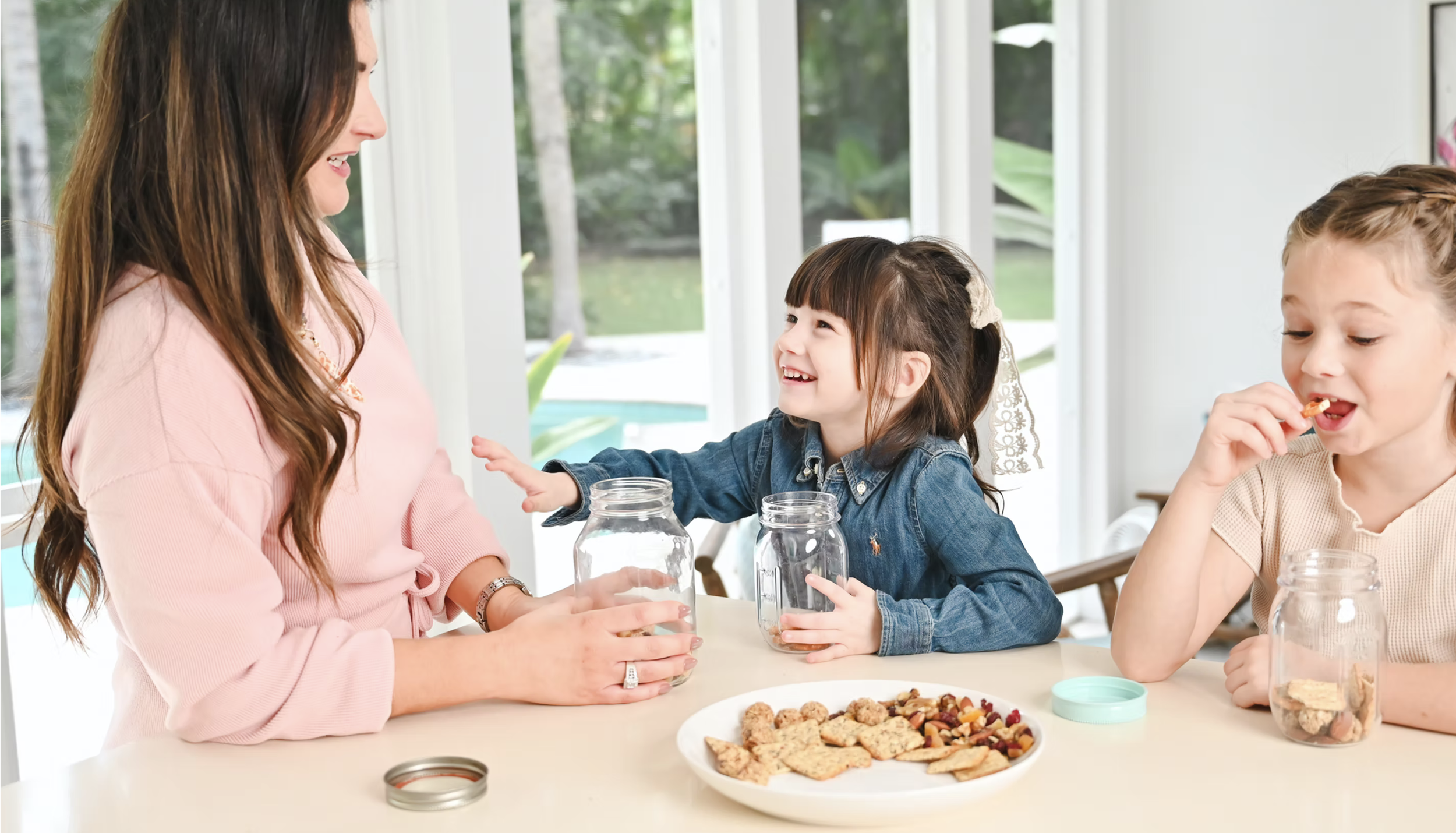 A woman and two young girls sitting at a table with jars and a plate of cookies and snacks, smiling and interacting in a bright, sunny room with large windows.