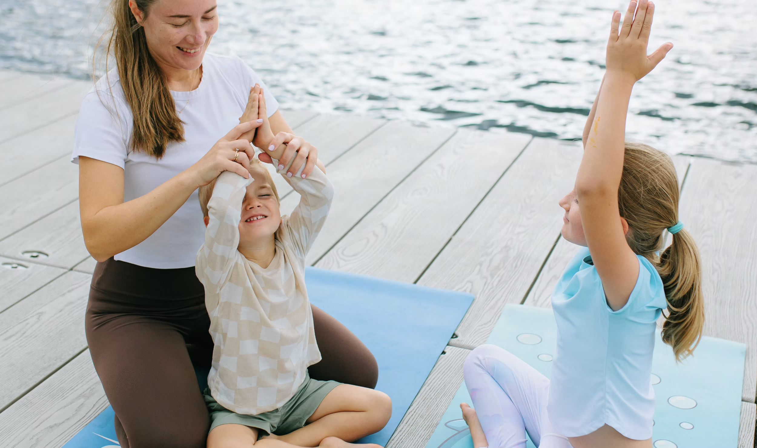 A woman helping a young boy with a yoga pose near a body of water, while a girl practices yoga and raises her arms, both on yoga mats on a wooden dock.