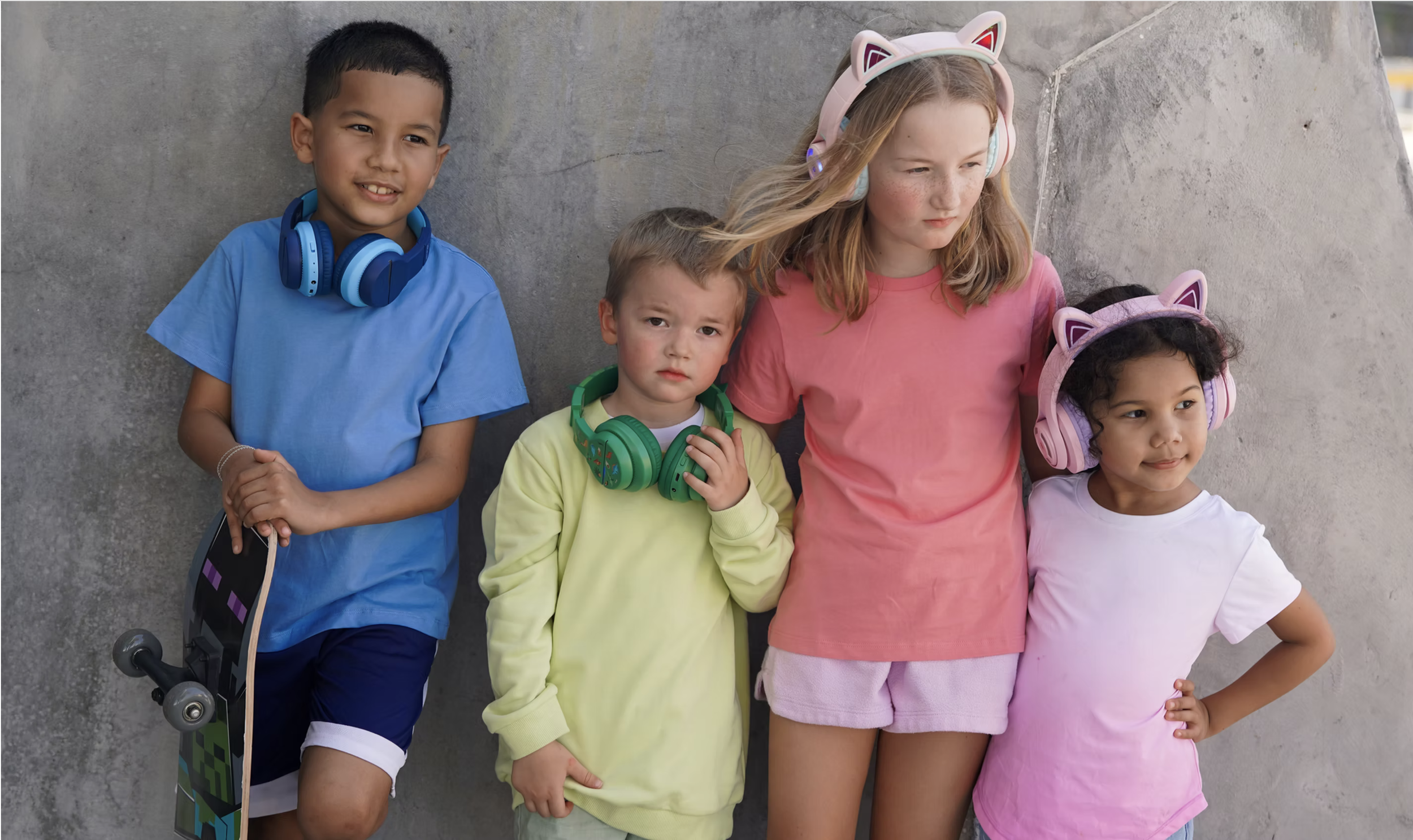 Four children standing against a grey concrete wall, wearing colorful clothes and headphones, with one holding a skateboard.