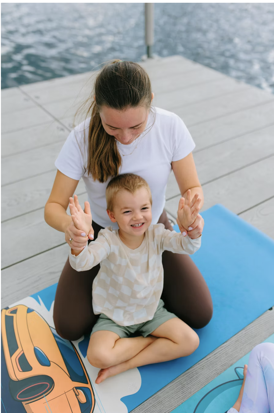 Woman and smiling child practicing yoga on a dock by the water, child sitting cross-legged on a yoga mat with a car illustration, woman kneeling behind, holding child's hands.