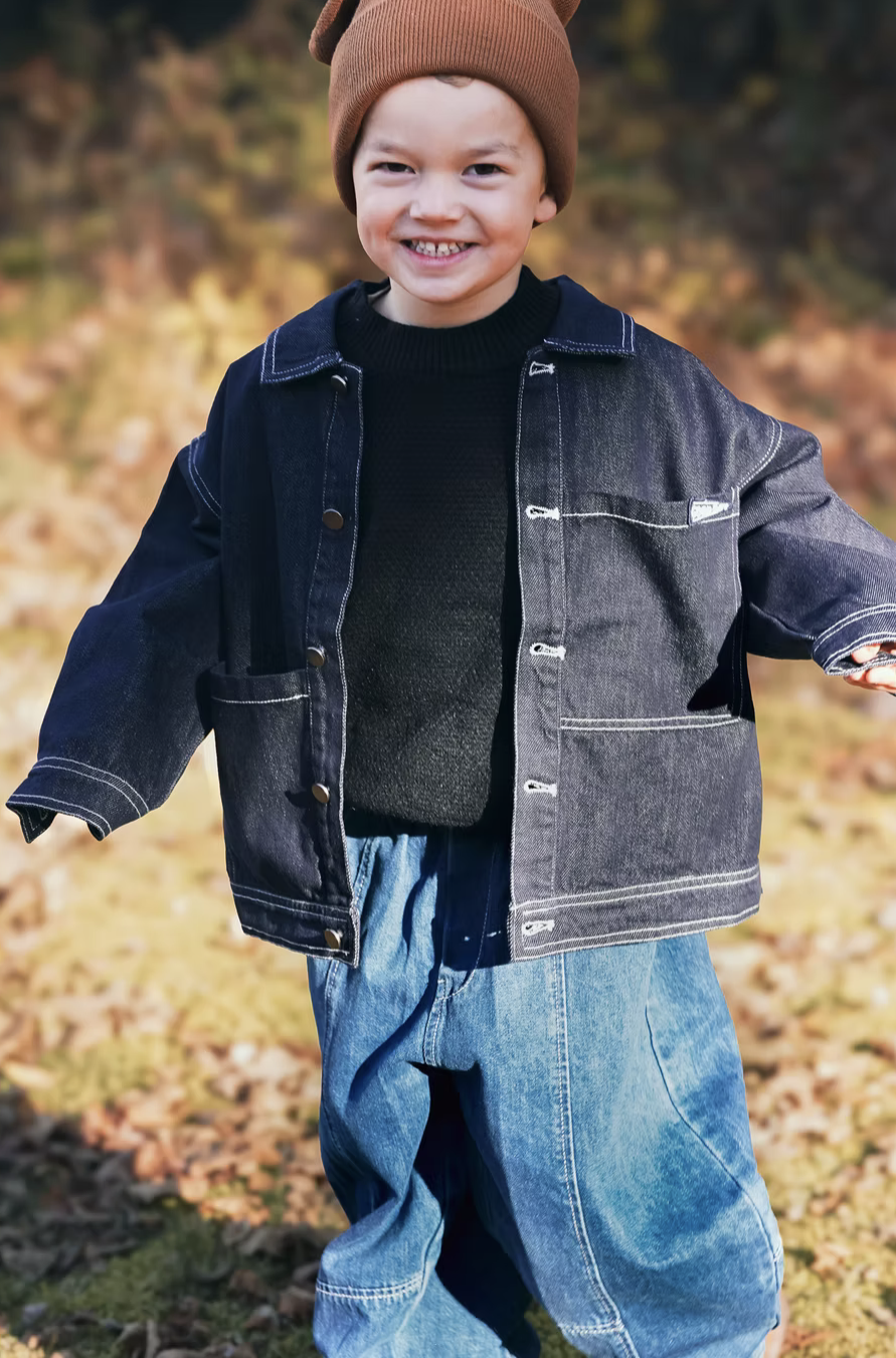 A young boy outdoors wearing a brown beanie, black sweater, denim jacket, and jeans, smiling at the camera.