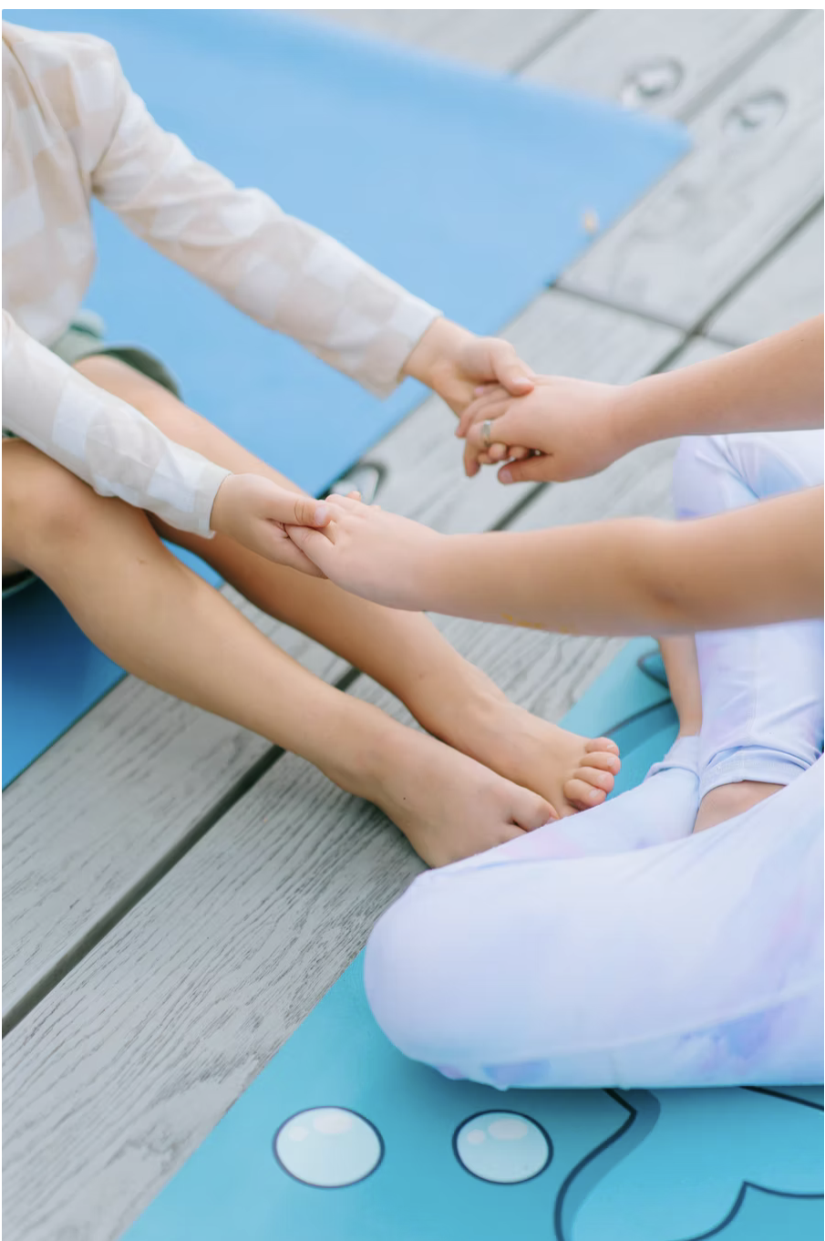 Two children sitting on outdoor wooden deck, holding hands, with yoga mats nearby.