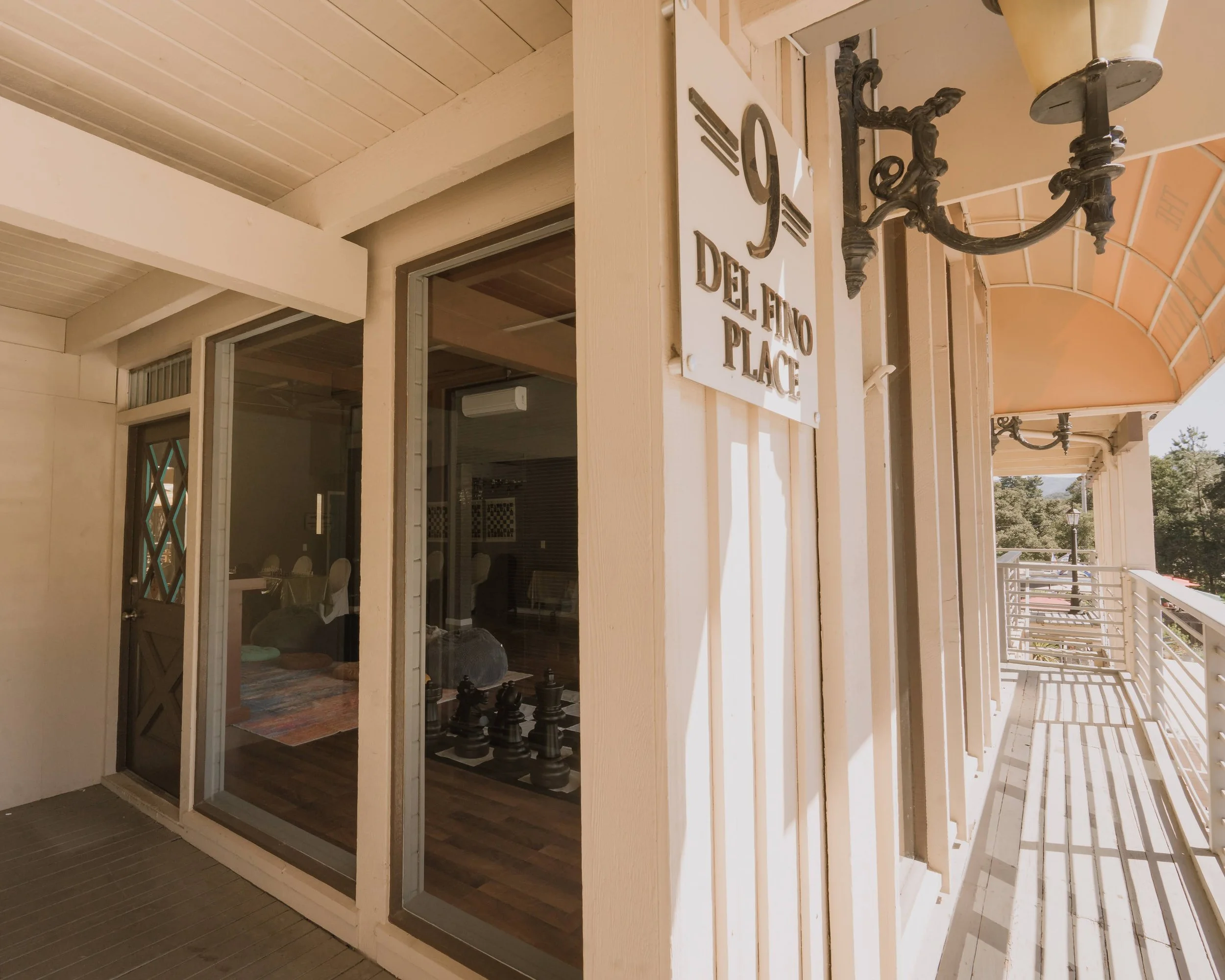 Exterior balcony of a building with a sign that reads '9 Del Fino Place,' showing a chess set inside and a scenic view of trees outside.