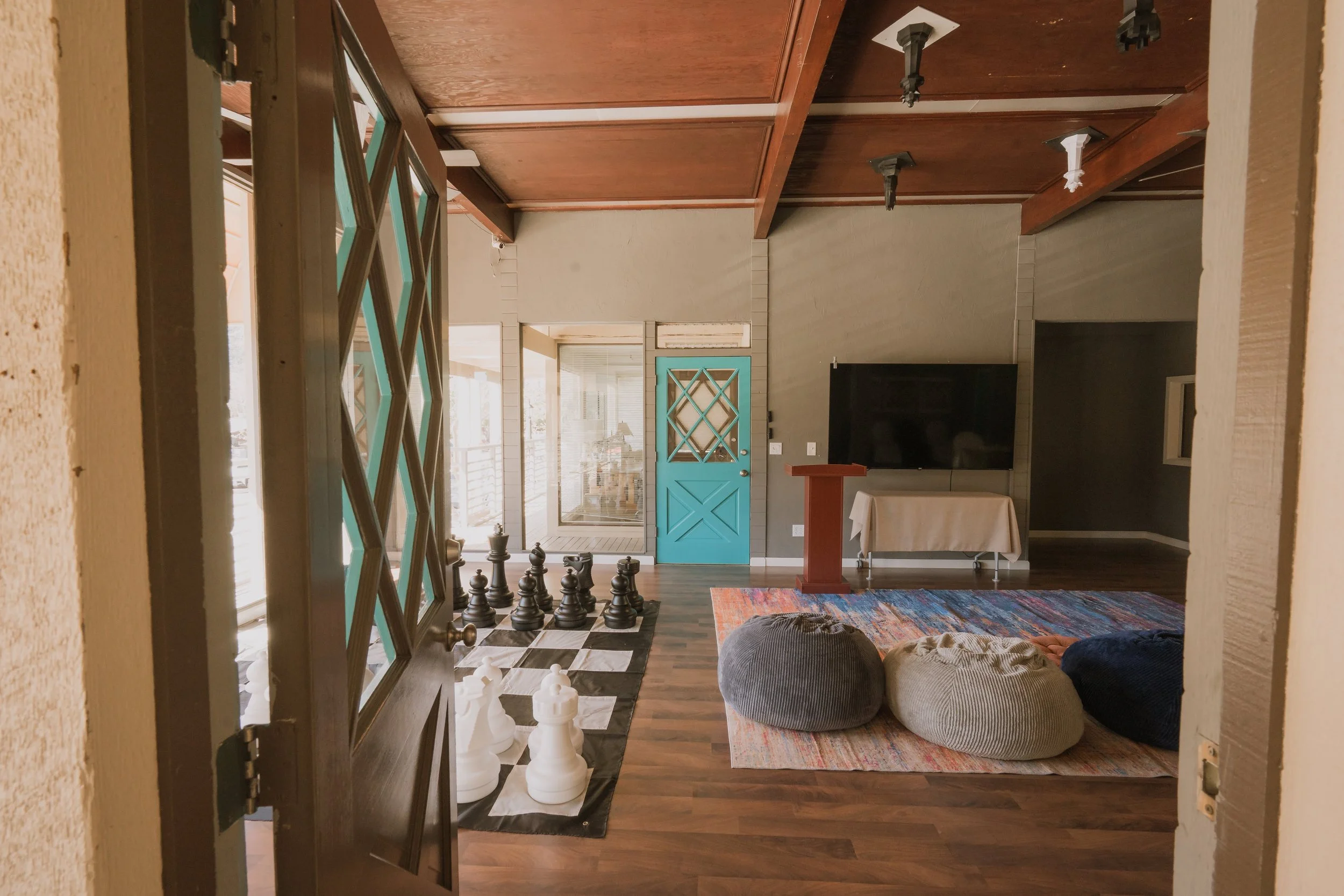 Room with a large black TV on the wall, a colorful area rug, three bean bag chairs, a chess set on the floor, a turquoise door, and windows with blinds.