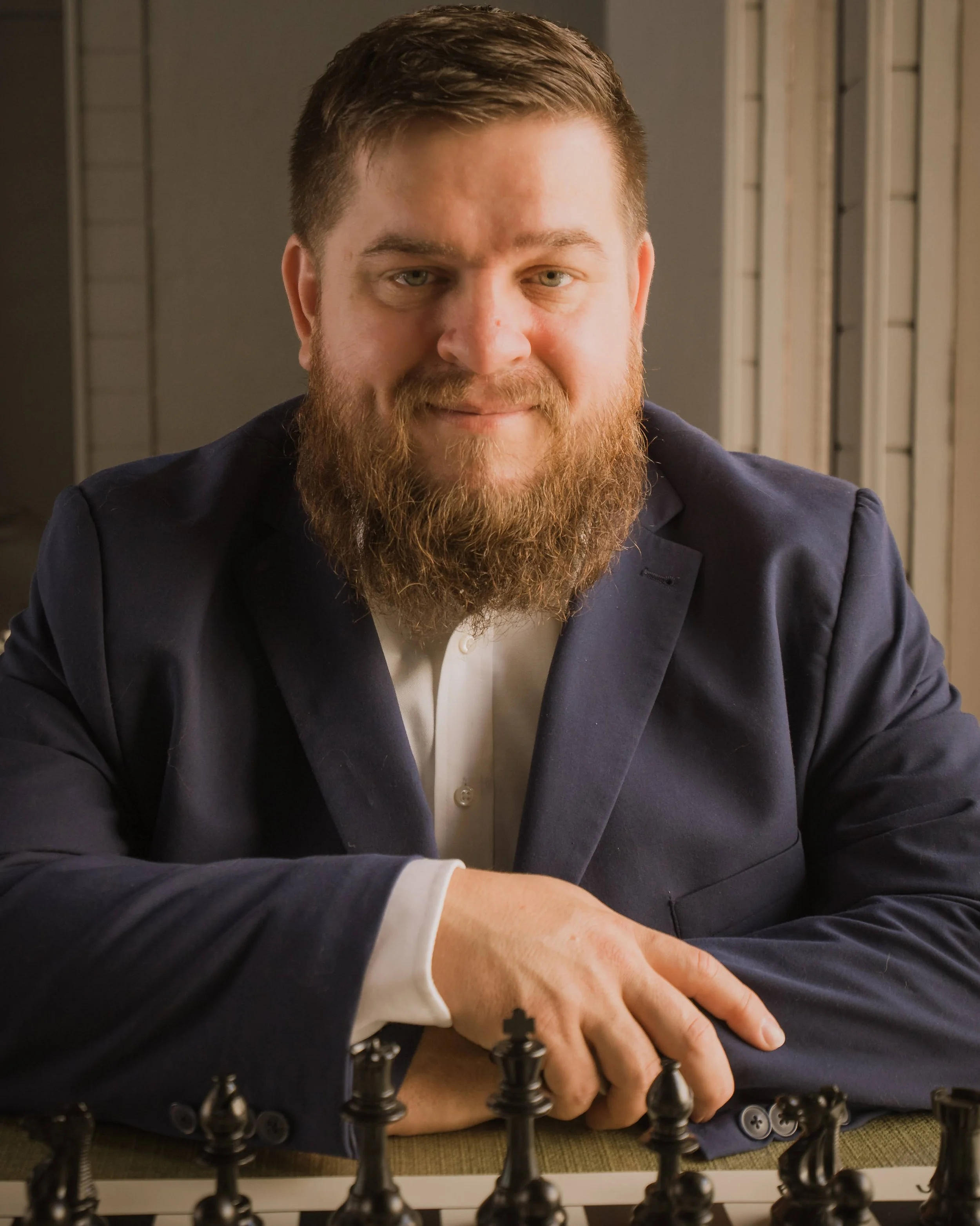 A man with a full beard and short hair, dressed in a dark suit and white shirt, sitting at a chessboard with a serious expression.