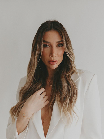 Portrait of a woman with wavy brown hair wearing a white blazer and a gold bracelet, posing against a plain background.
