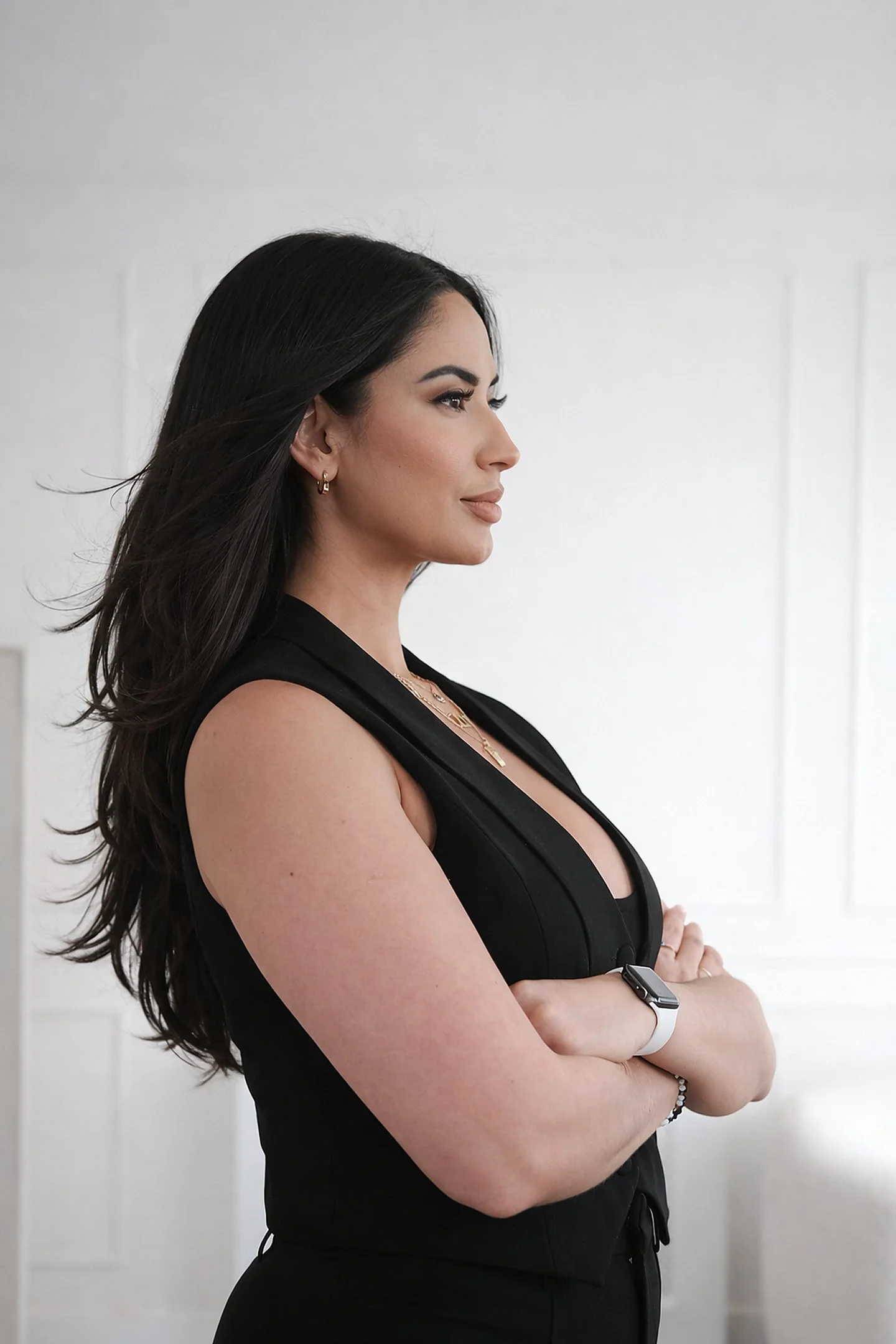 Profile of a woman with long dark hair, arms crossed, wearing a black sleeveless dress, gold jewelry, and a smartwatch, standing against a white wall.