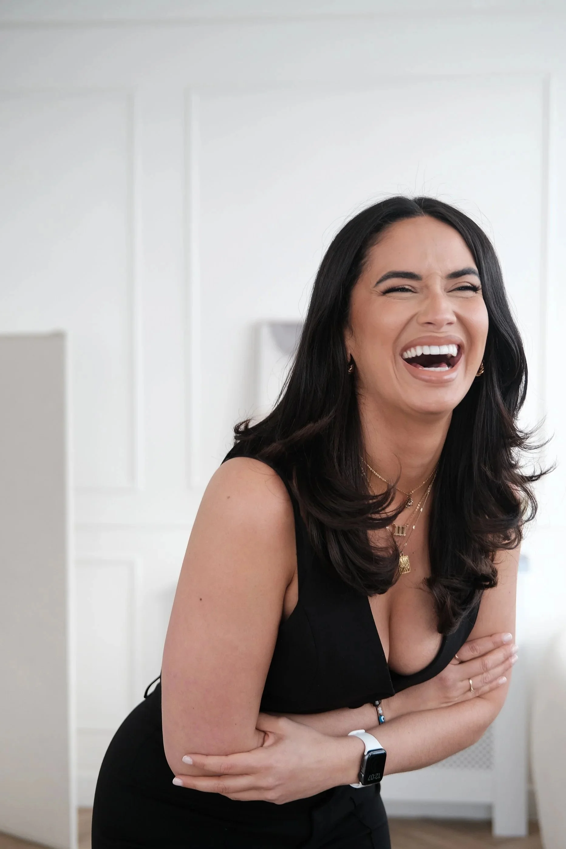 A woman with long dark hair, wearing a black sleeveless top, is laughing with her arms crossed, showing her teeth and her eyes closed.