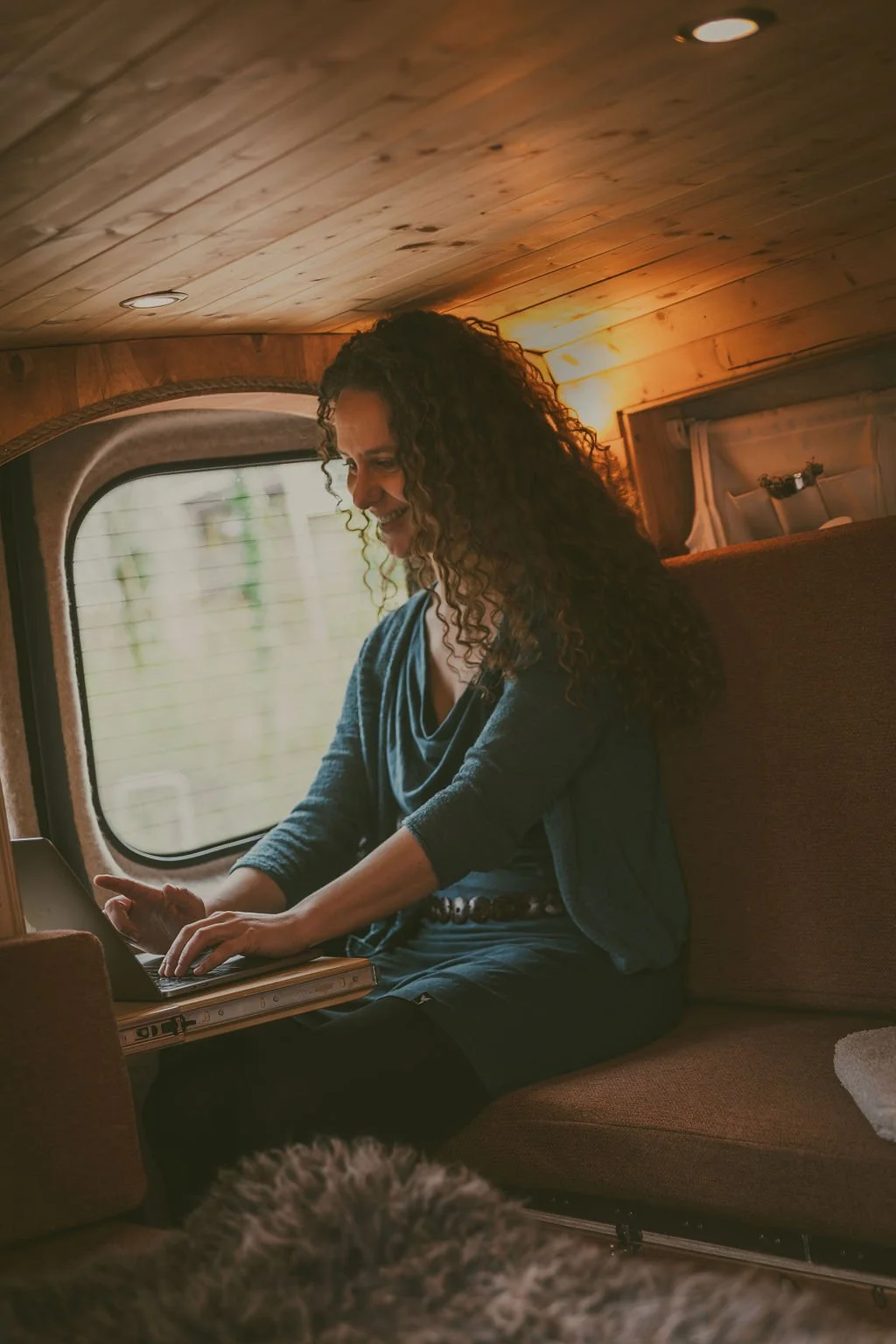 een vrouw met lang, krullend haar die op een trein zit, terwijl ze op een laptop werkt, met een houten interieur en een raam op de achtergrond.