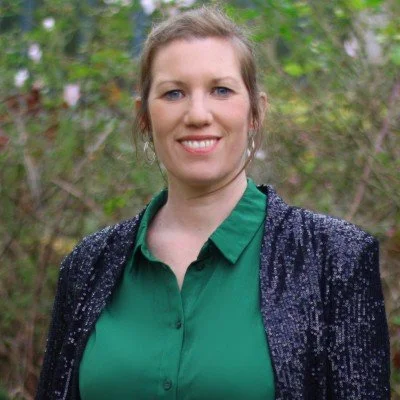 Vrouw die buiten poseert met groene blouse en zwarte glittervest, omgeven door groene bomen en struiken.