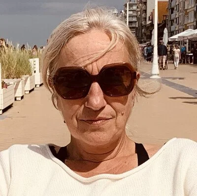 Een oudere vrouw met roze haar, grote donkere zonnebril en een witte trui die selfie neemt op een promenade met winkels en mensen op de achtergrond.