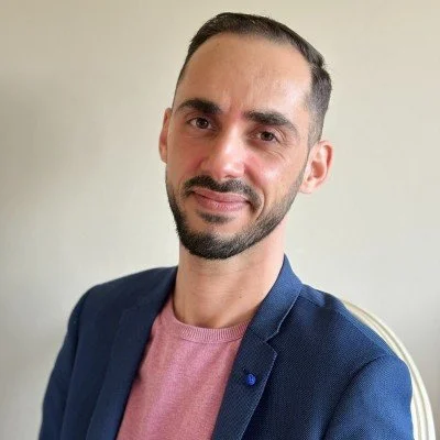 Portret van een man met een baard, in een blauwe blazer en een roze trui, zittend op een stoel tegen een lichte muur.