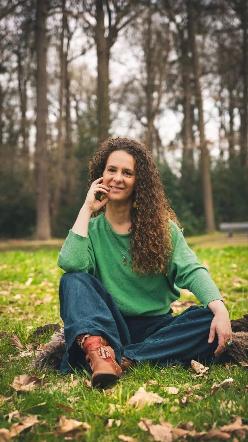 Vrouw met lang, krullend haar in een groene trui zittend op gras in een bos