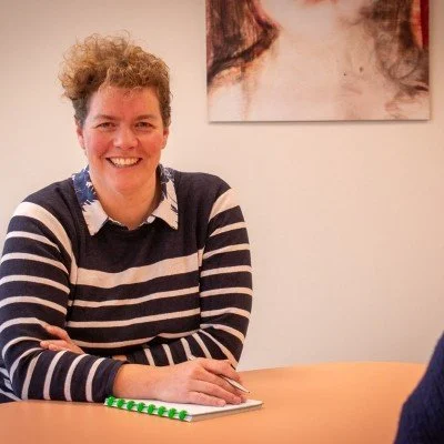 Vrouw met kort, krullend haar en een strepentrui, lachend tijdens een gesprek in een kamer.