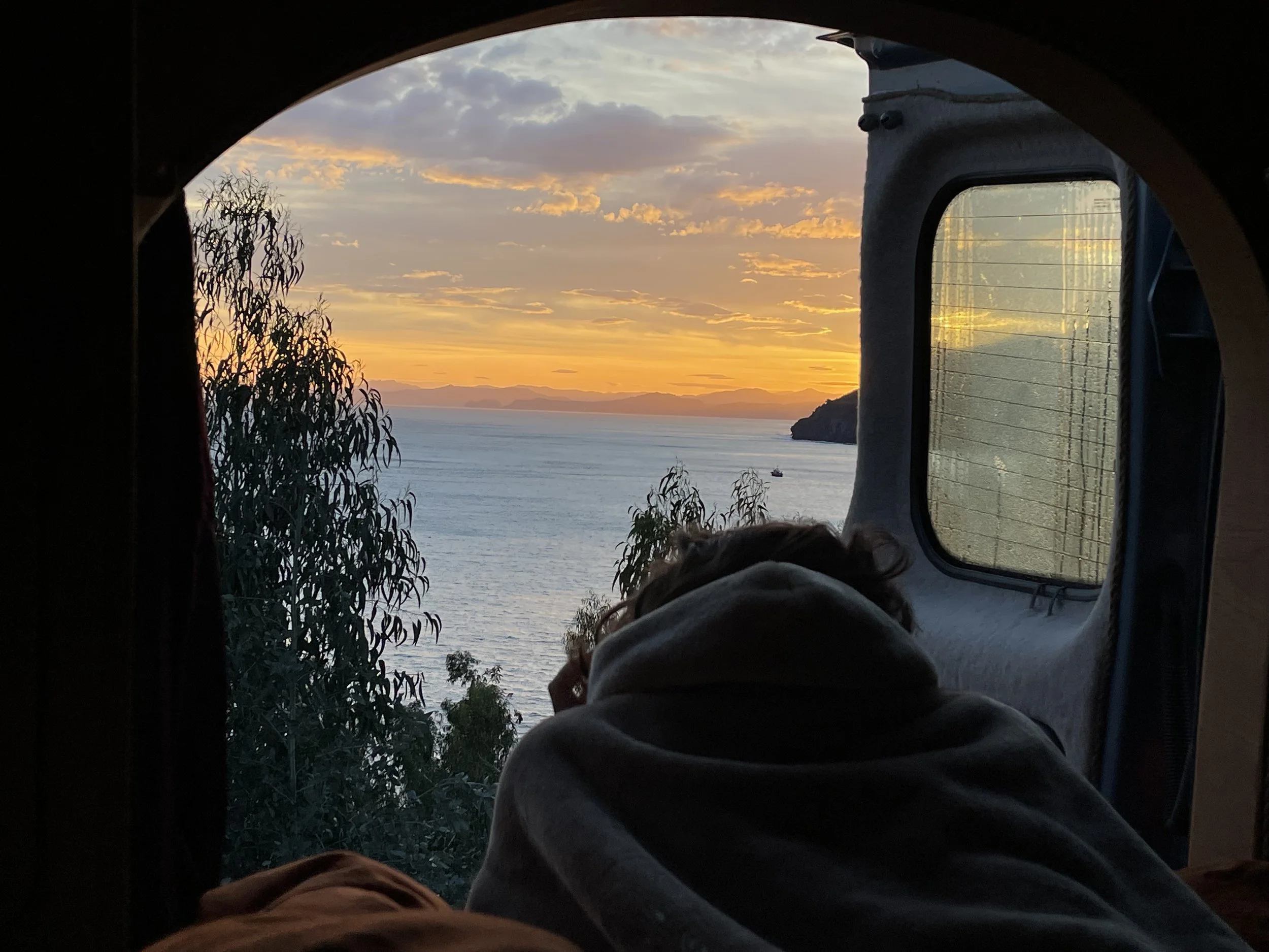 Binnenkijkend vanuit een camper of bus, met een persoon die in bed ligt, uitkijkend op een zee en een zonsondergang met gekleurde wolken, omringd door bomen en een eiland in de verte.