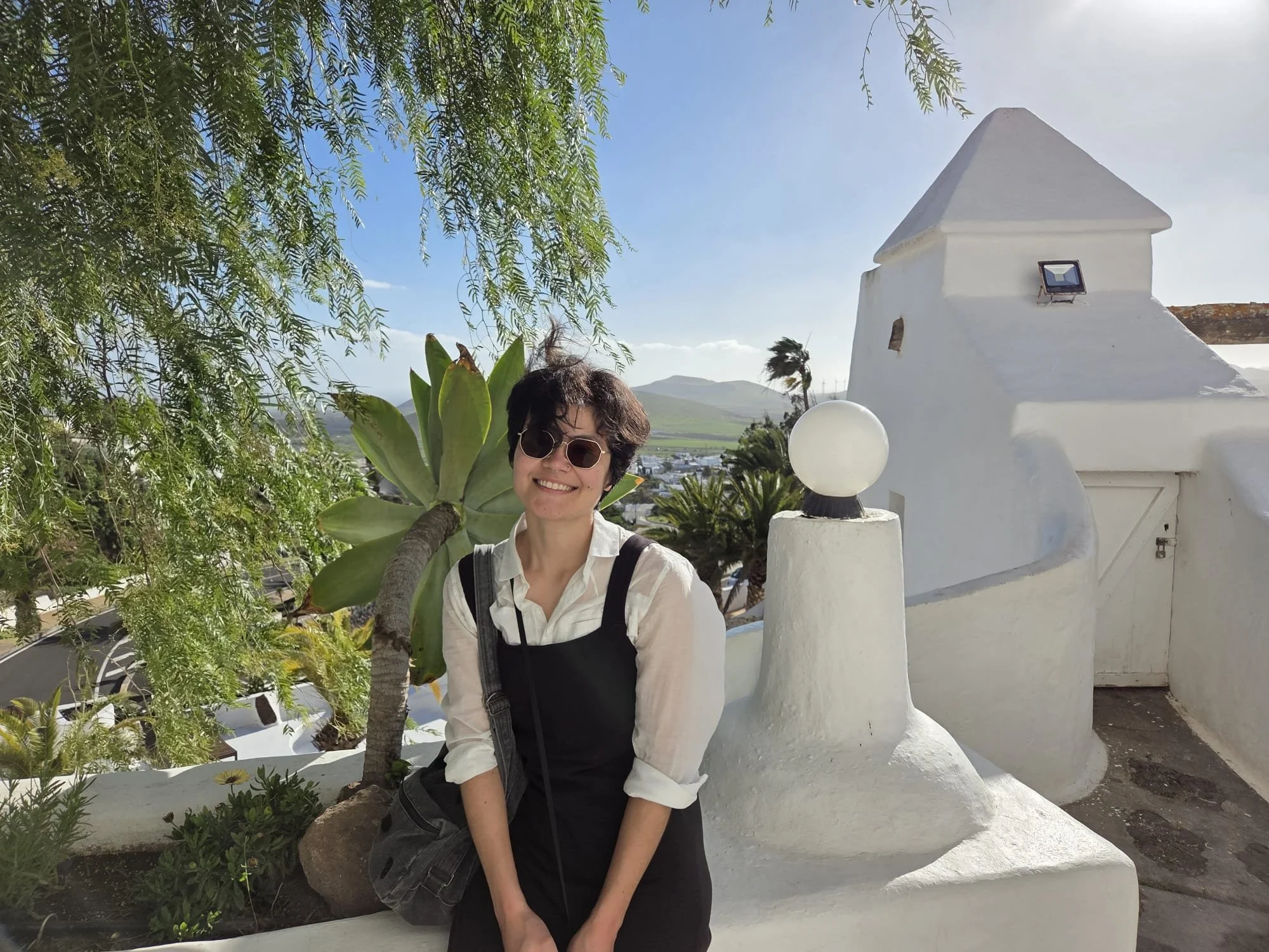 A smiling woman with short dark hair, sunglasses, and a black dress over a white shirt standing in front of white stucco architecture and green plants, with mountains and a clear blue sky in the background.