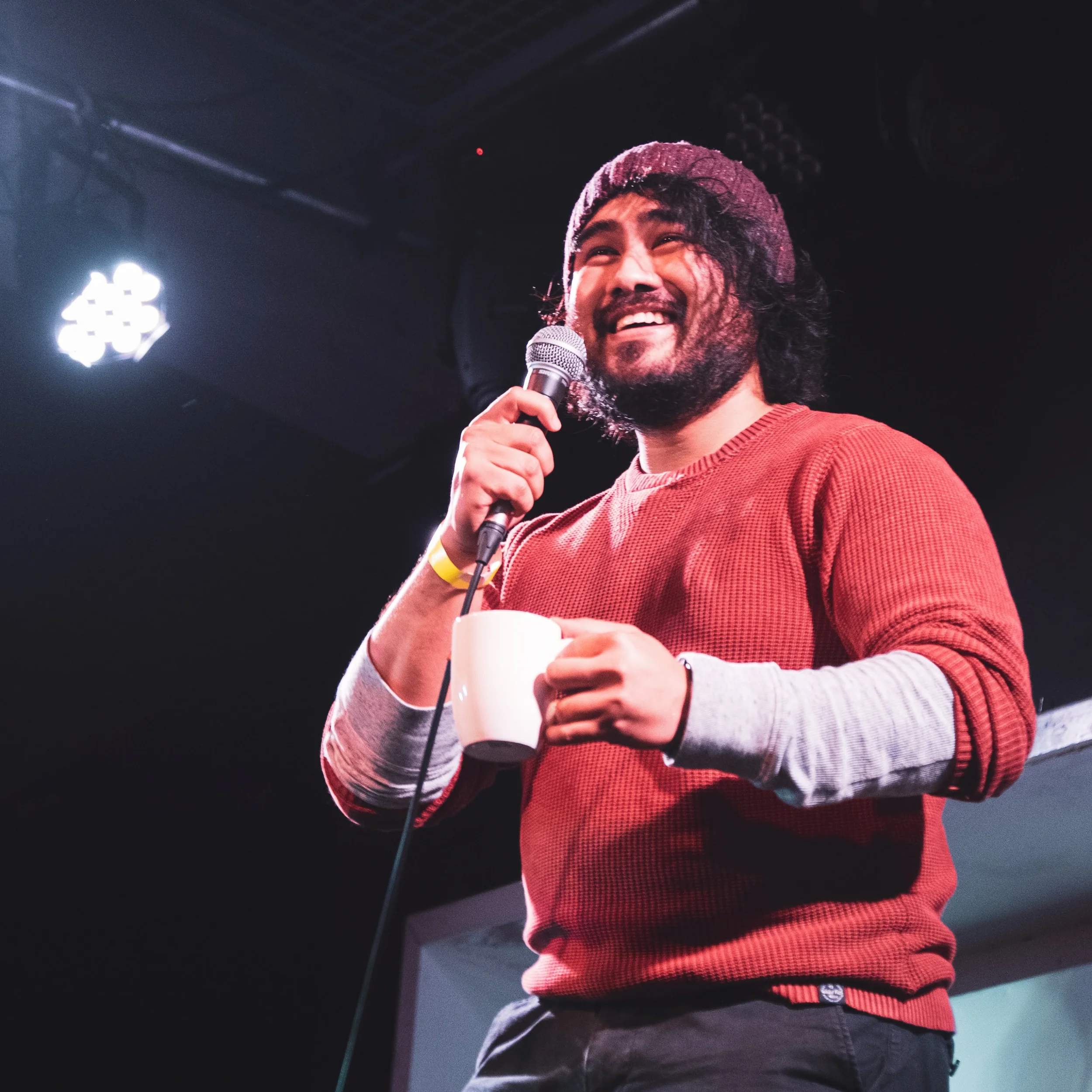 A man with a beard, wearing a beanie and a red sweater, smiling while holding a white mug and speaking into a microphone on stage.