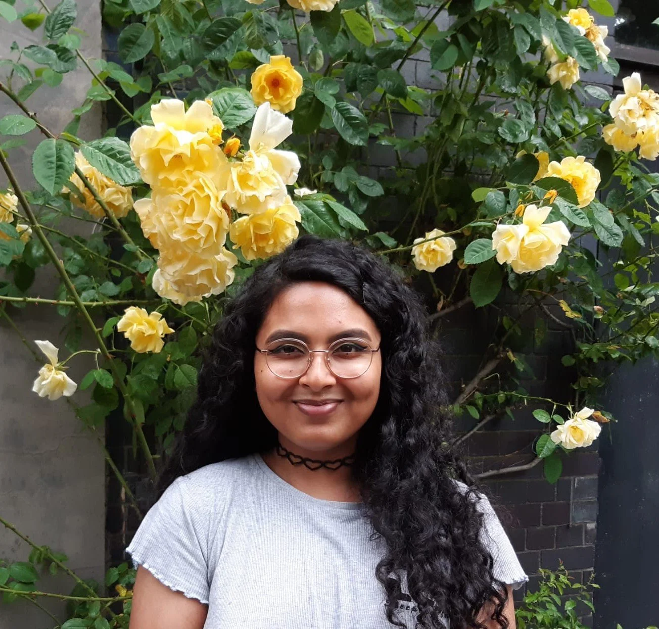 Young woman with glasses and long curly hair smiling in front of a large yellow rose bush with green leaves.